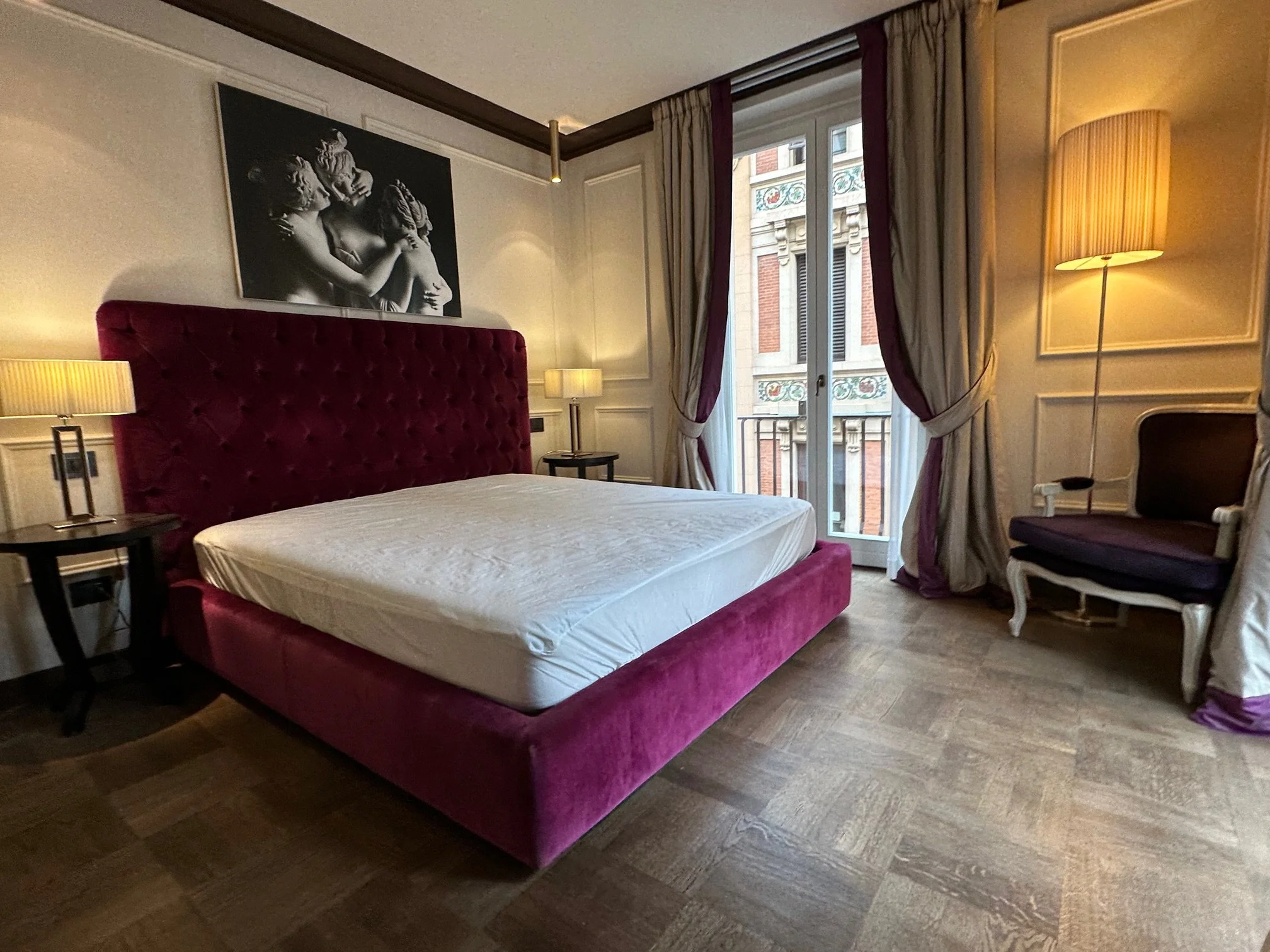 A hotel room with a large plush red velvet bed, bedside lamps, a window with ornate curtains, and a black and white artwork of classical sculptures on the wall.