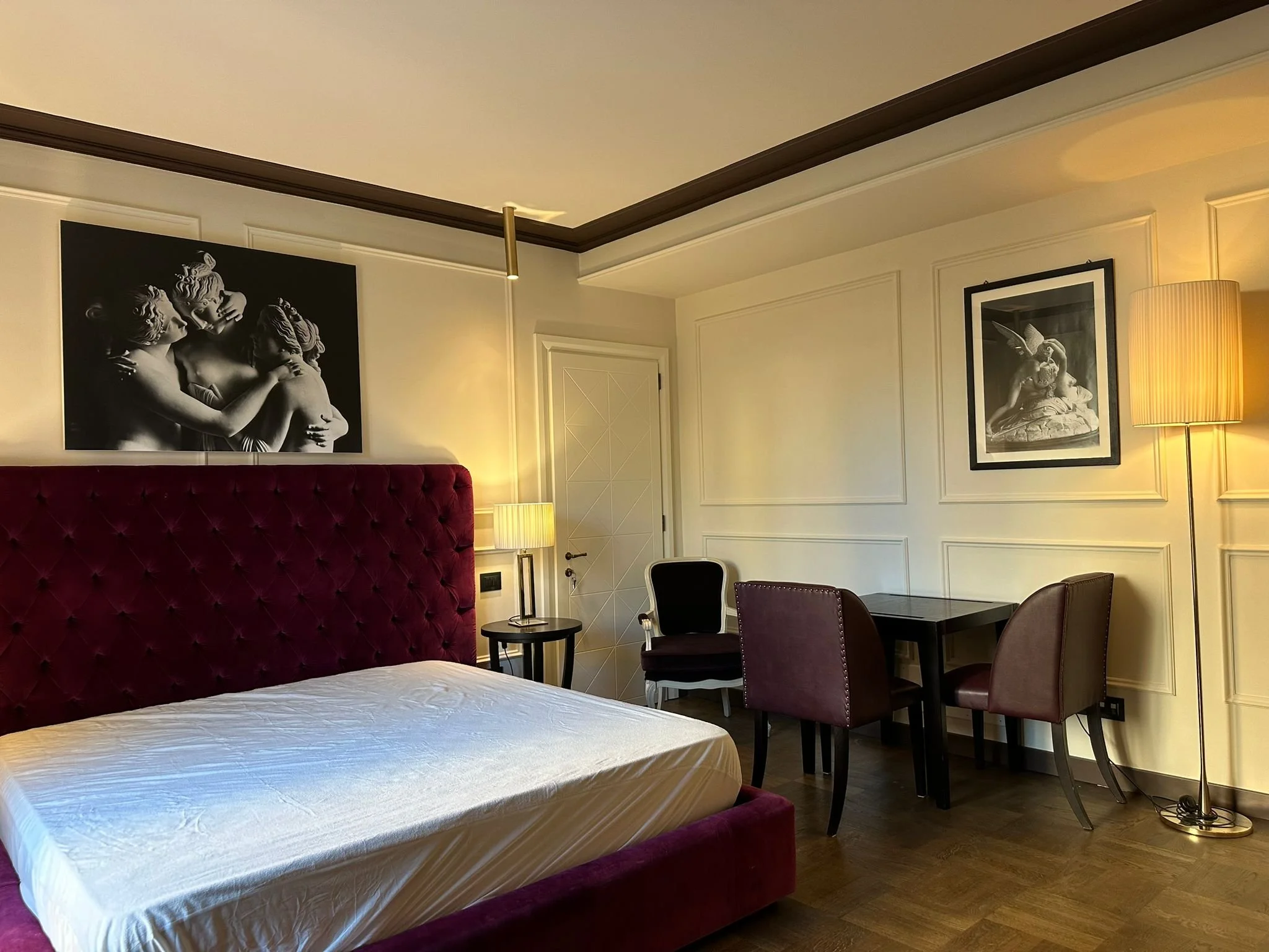 A hotel room with a bed featuring a deep red, tufted headboard, two black and white framed photographs on the wall, a small round side table, a black chair, a dining table with purple upholstered chairs, and a tall floor lamp in a corner.