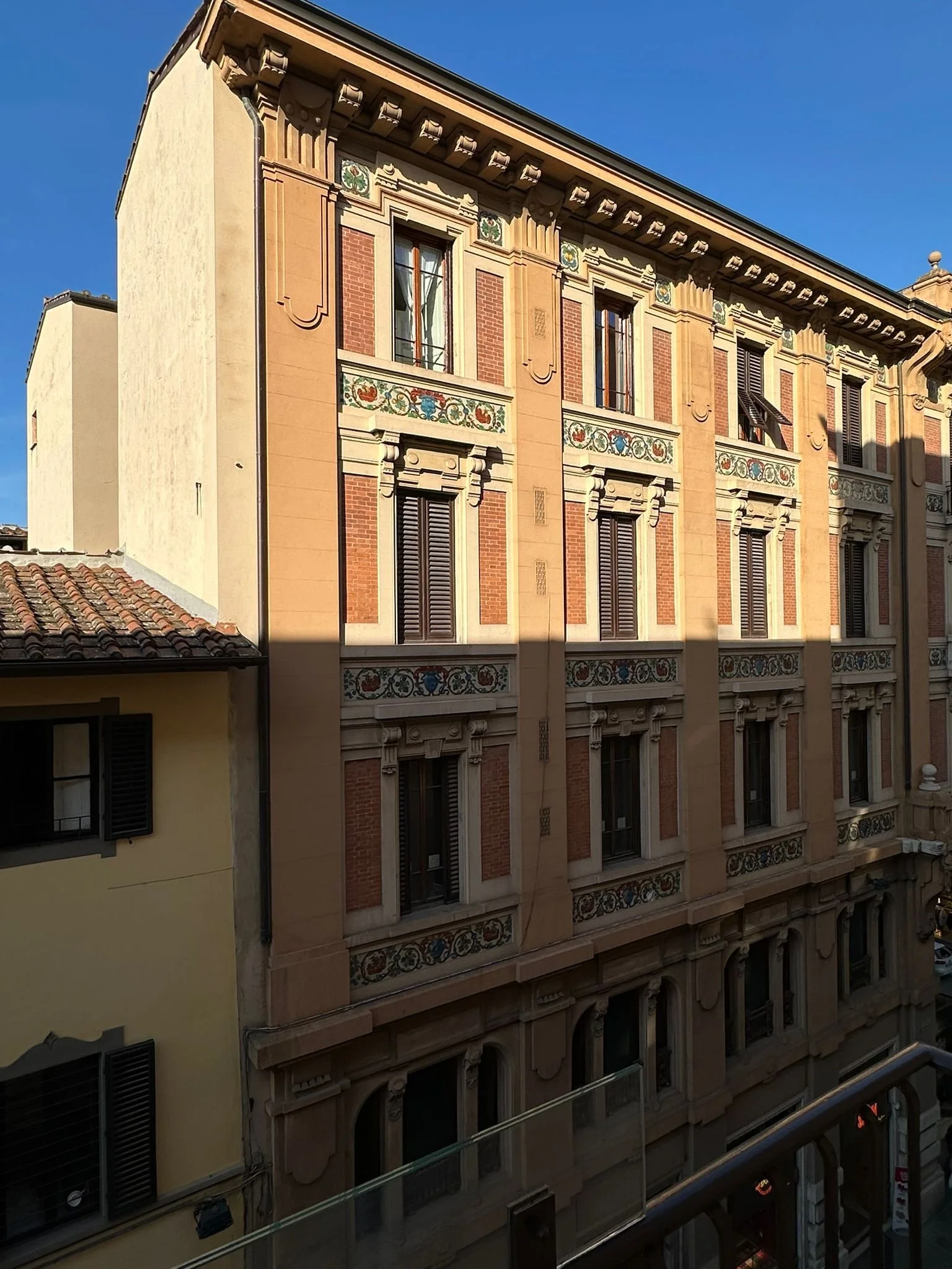 View of a historic building with ornate architecture, multiple windows with shutters, decorative floral and geometric patterns, and a small rooftop balcony under a clear blue sky.