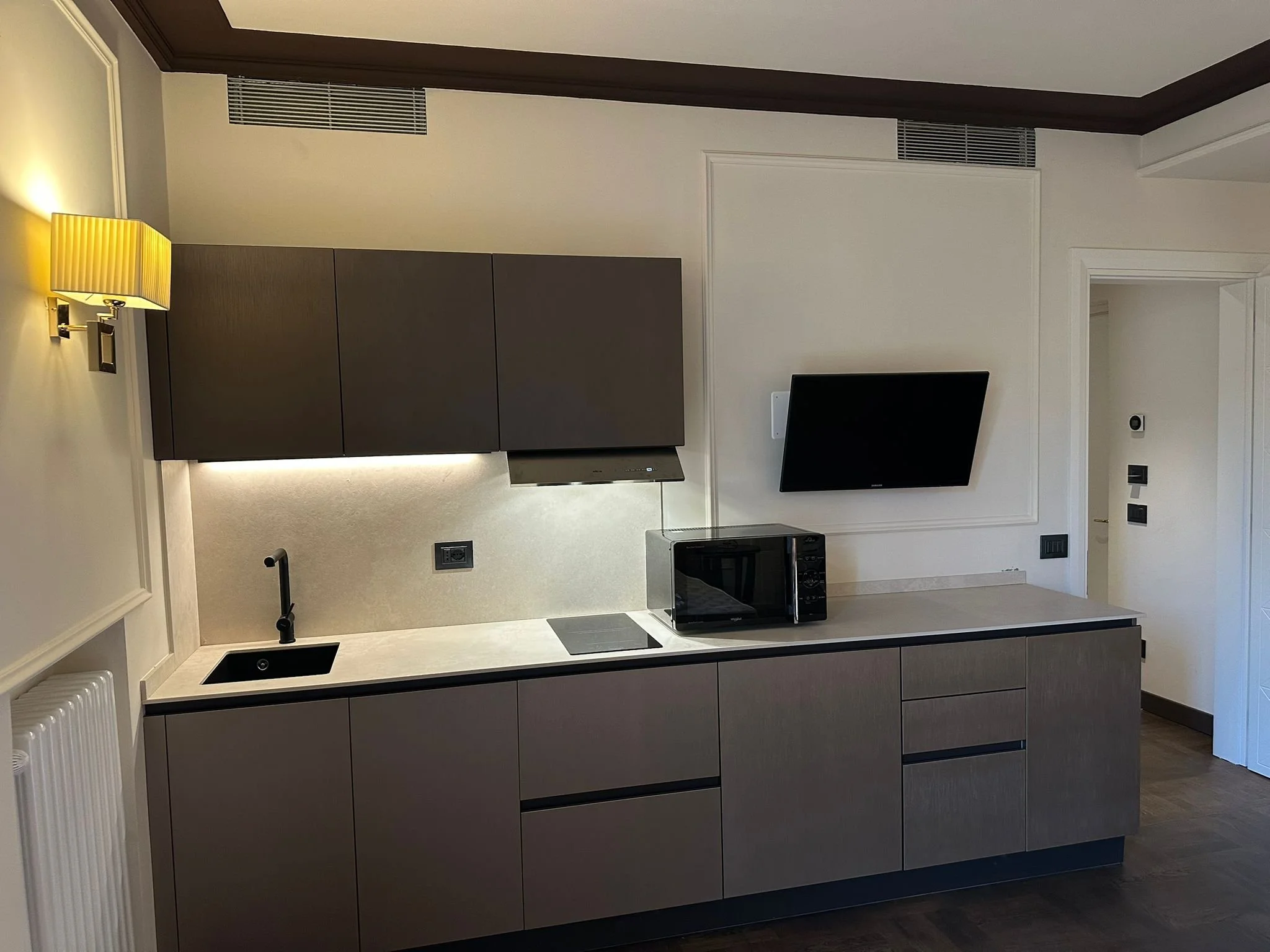 Modern kitchen with dark cabinets, a small black sink, a black microwave, and a wall-mounted TV. The room features white walls with dark wood trim and flooring.