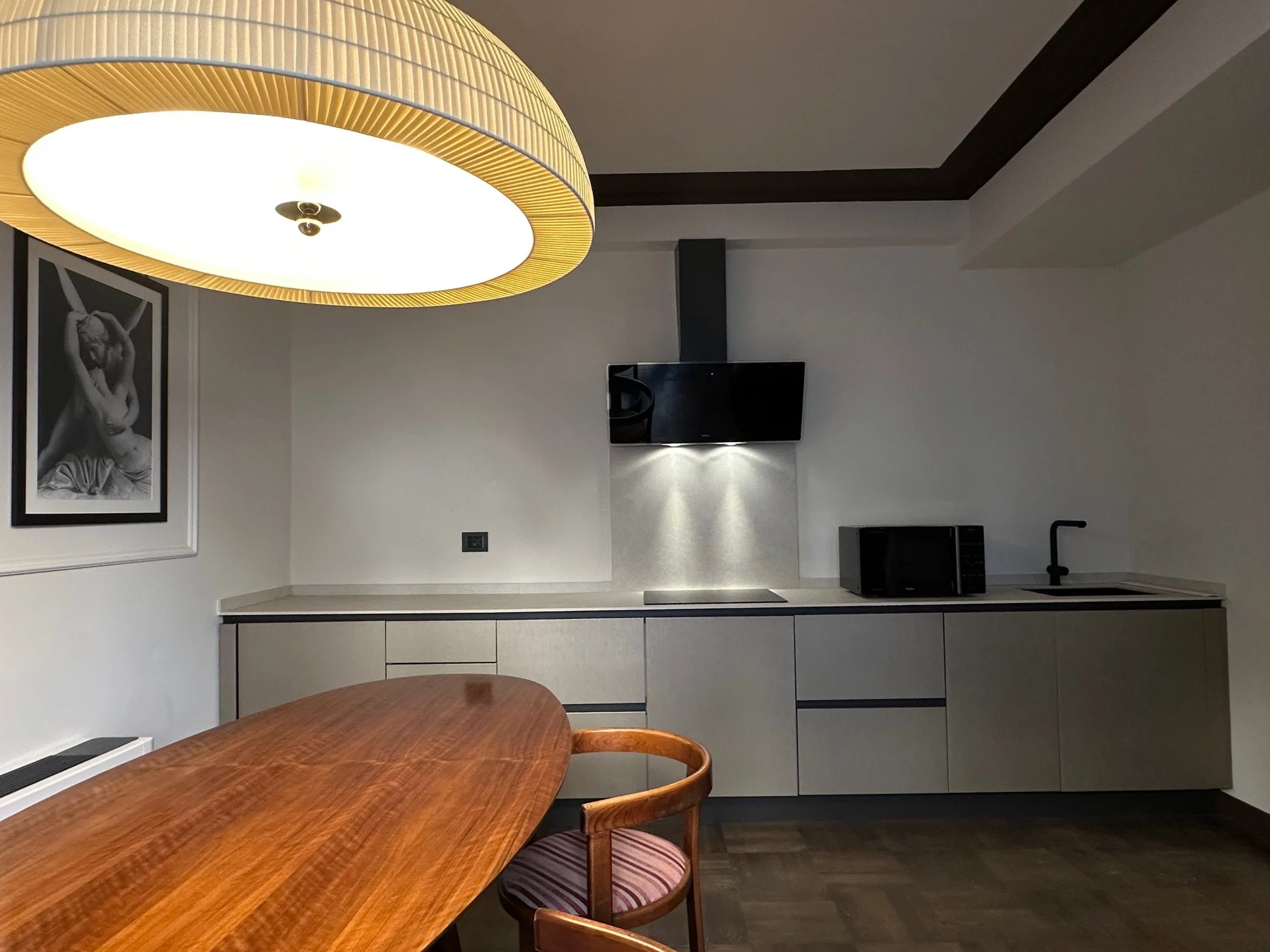Modern interior room with a round wooden dining table, wooden chairs, a black flat-screen TV on the wall, black and white framed photo of a woman, and a ceiling light fixture.