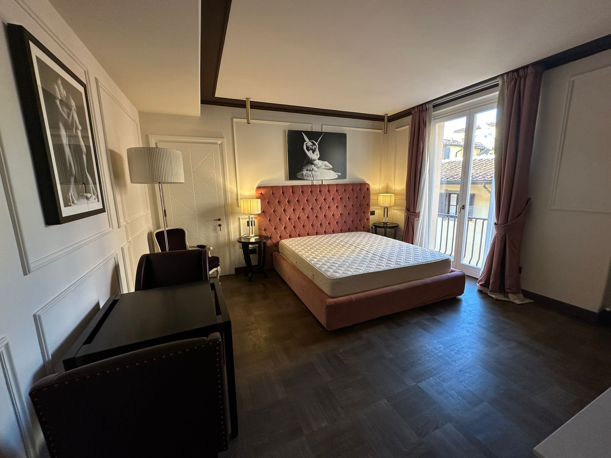 A hotel or bedroom with a bed, pink headboard, two bedside lamps, framed black-and-white art, and a large window with curtains showing rooftops outside.