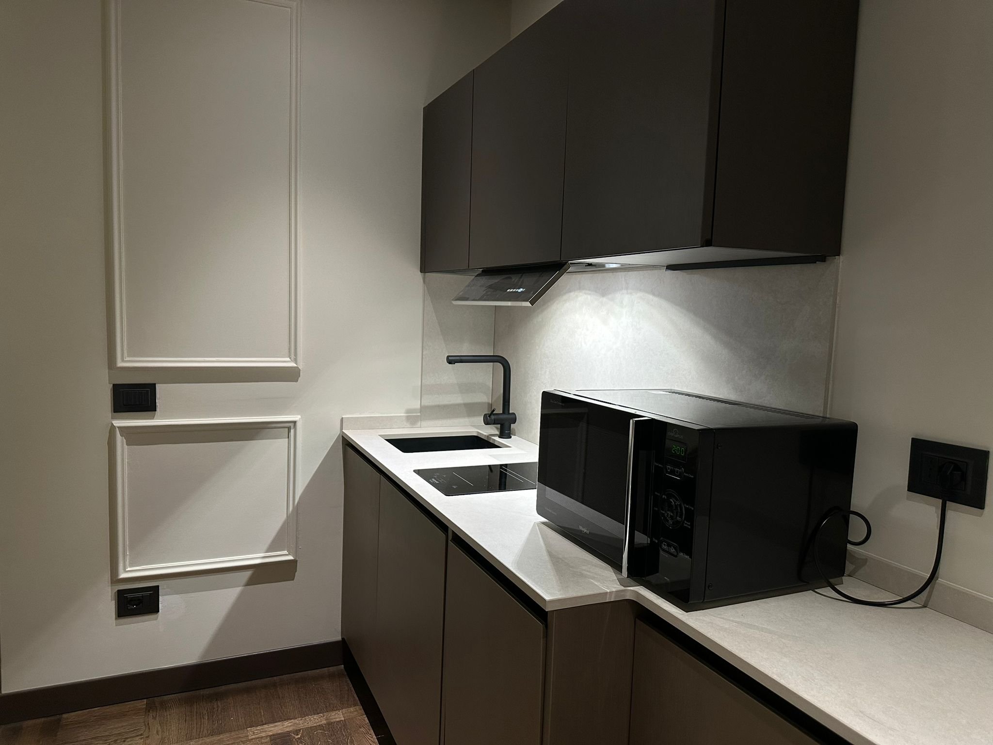 Modern kitchen with dark cabinet doors, a white countertop, black sink with a black faucet, a black microwave oven, and a wall with white paneling and electrical outlets.