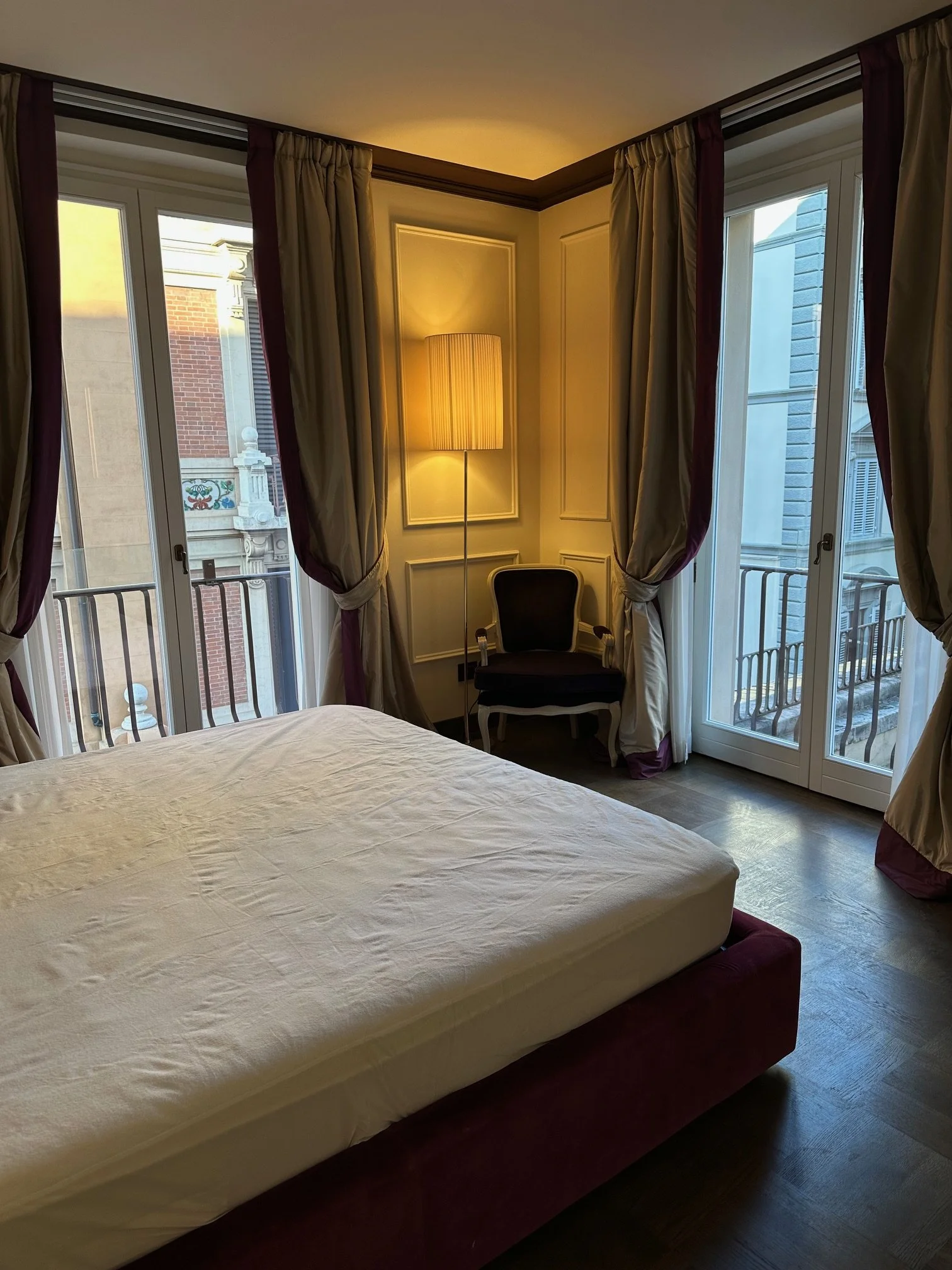 A hotel room with a large bed, tall windows with curtains, a chair, and a tall lamp in a corner