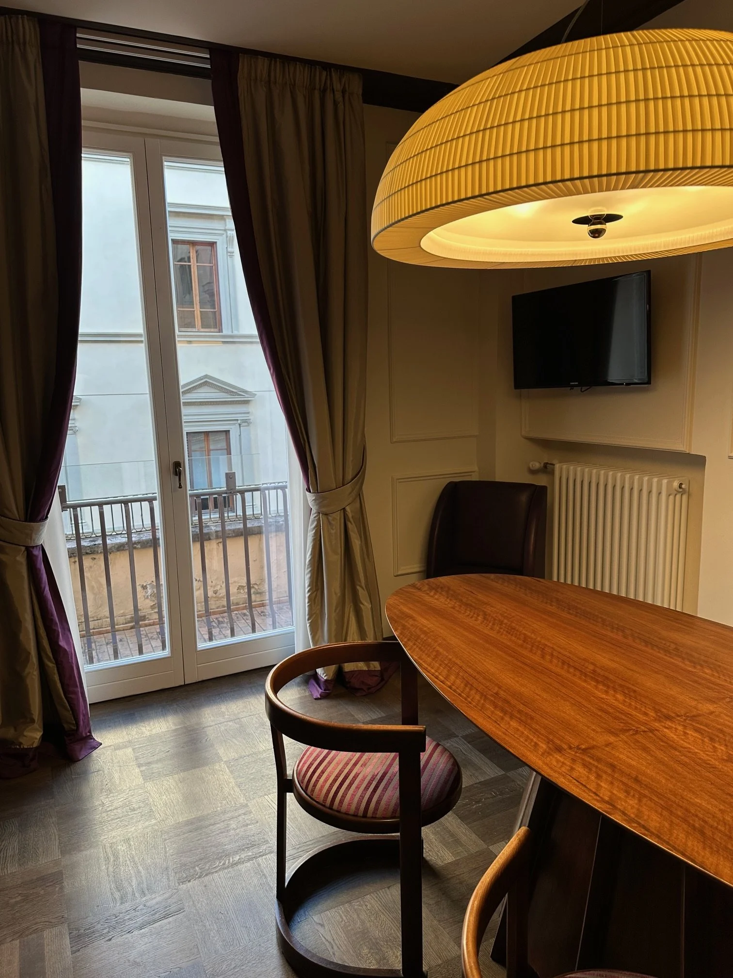 Interior of a room with a round wooden dining table, a chair with a striped cushion, a flat-screen TV on the wall, large windows with purple curtains, and a large yellow ceiling lamp.