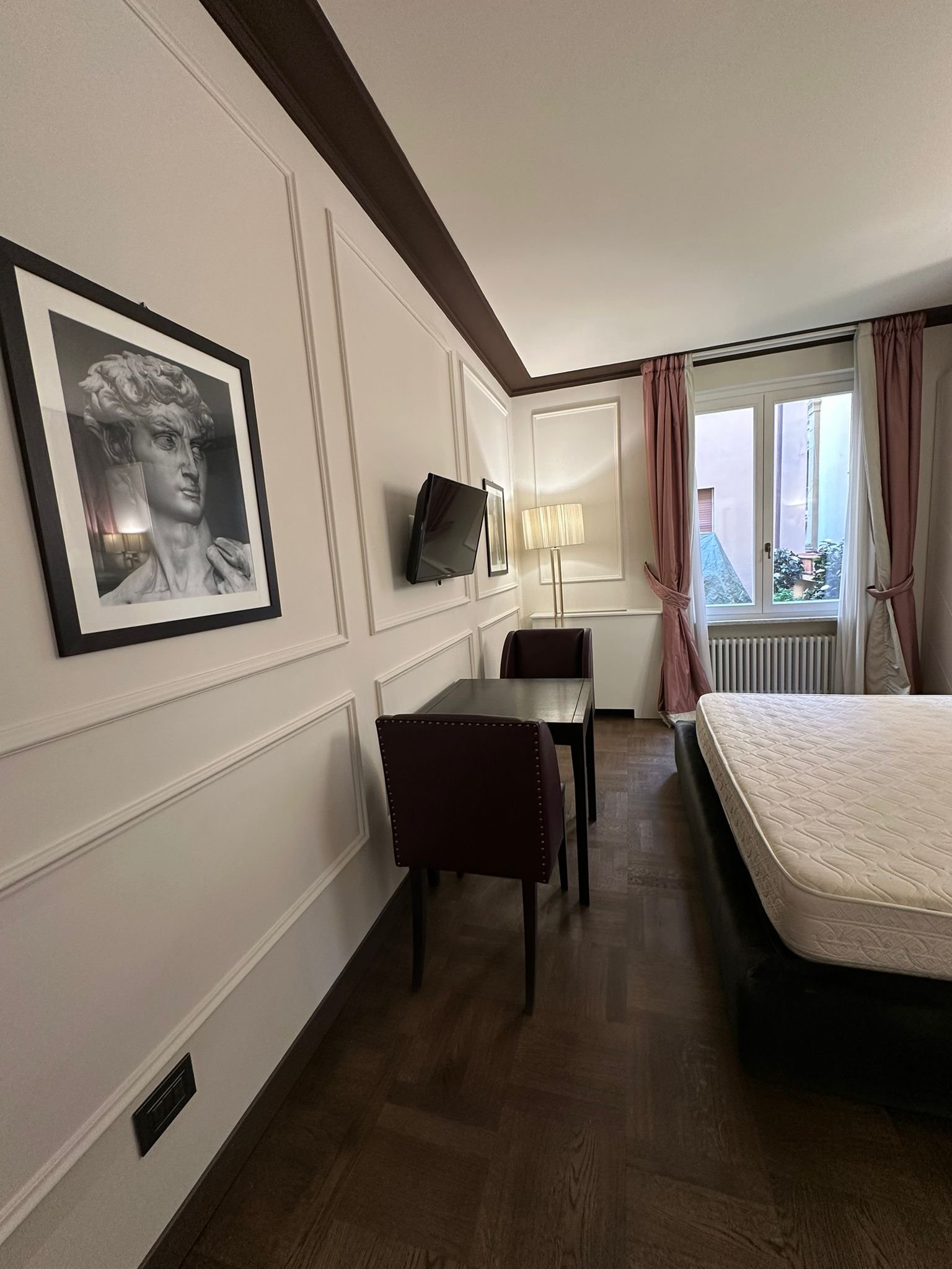 A corner of a hotel room with a bed, a small desk with a dark red chair, a lamp, a framed black-and-white portrait on the wall, and a window with pink and white curtains.
