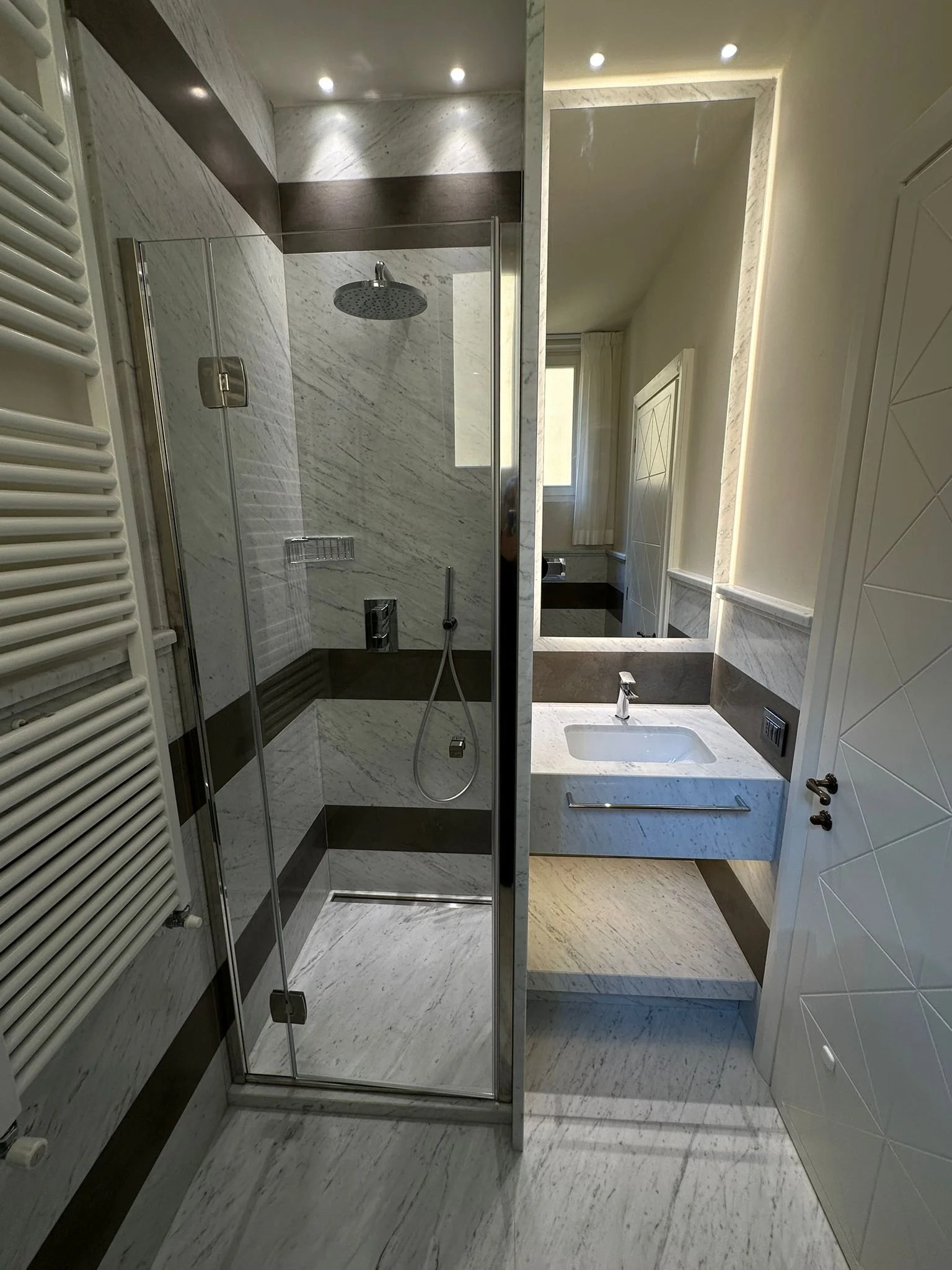Modern bathroom with marble floors and walls, walk-in shower with glass doors, a small sink with a mirror, and a window with curtains.