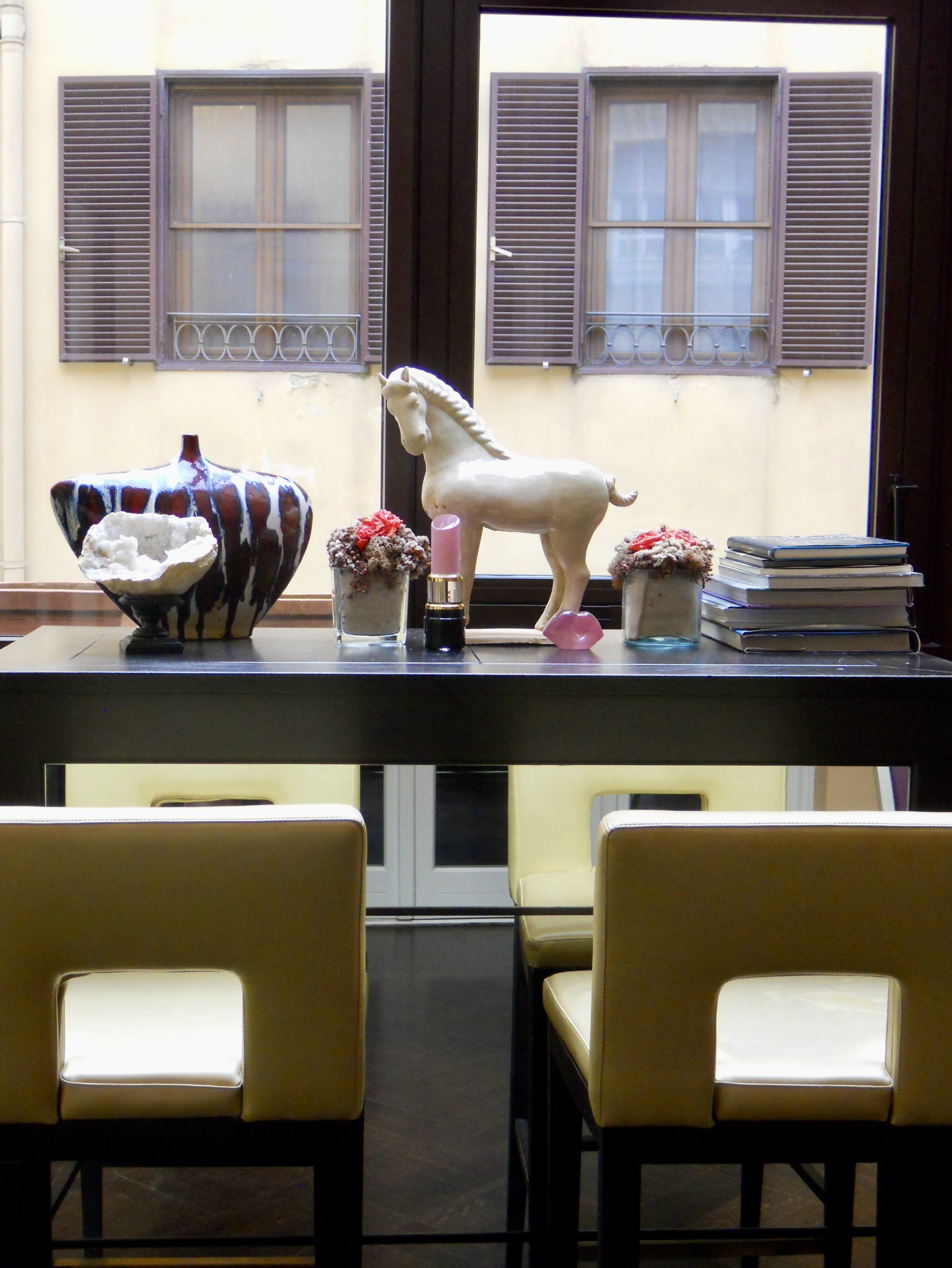 View of a table with a decorative horse figurine, two potted plants, a pink glass object, a stack of books, and a candle, set in front of a window with purple shutters and a yellow wall outside.