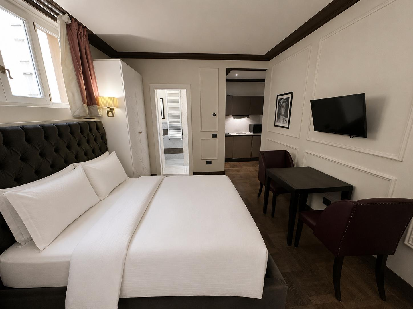 A hotel room with a large bed, white bed linens, and a tufted headboard. There are two pillows on the bed. Next to the bed, on the wall, is a yellow wall lamp. A window with curtains is above the bed. In the corner, a wardrobe is visible. A small tab