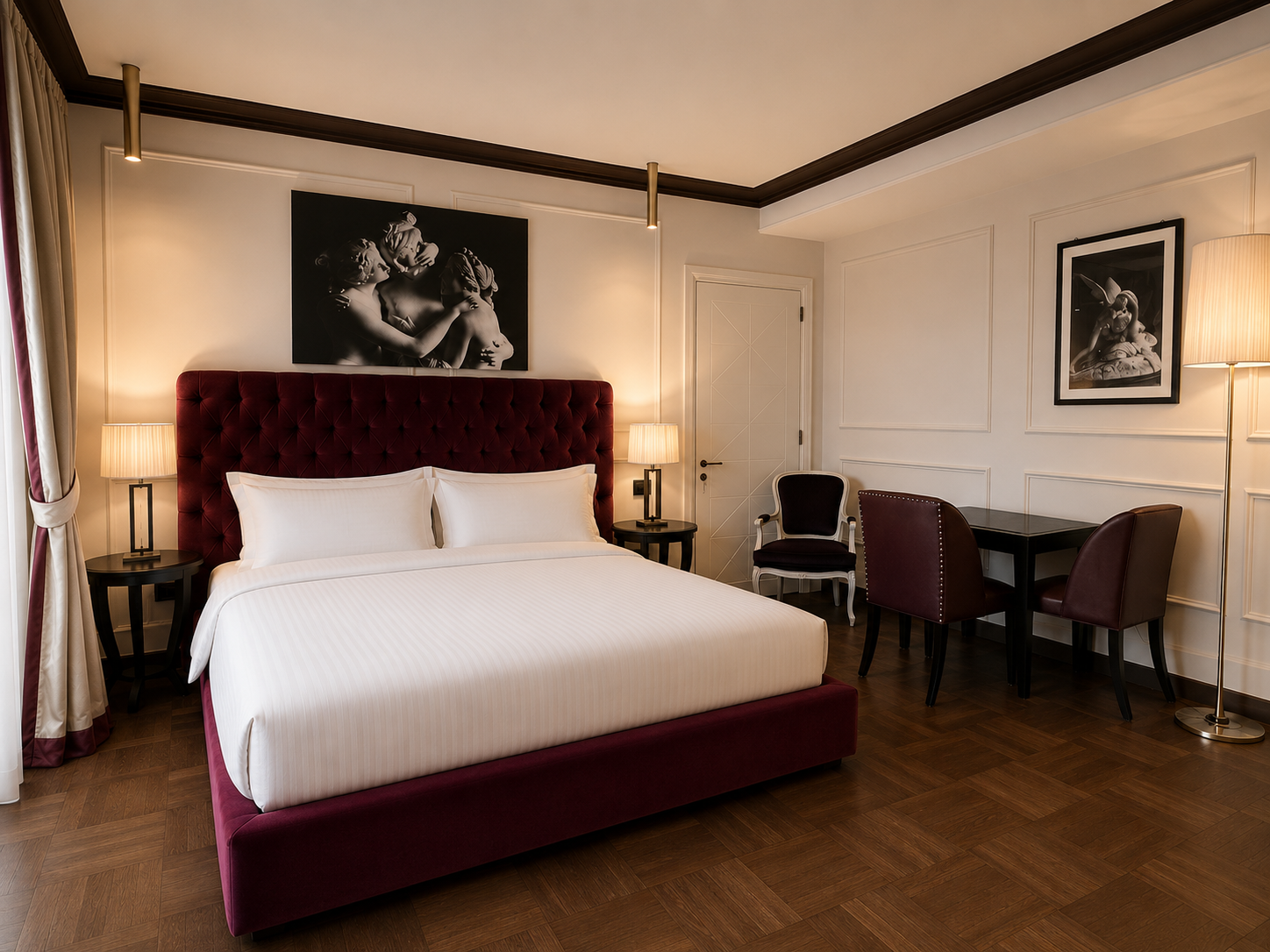 Hotel bedroom with a large bed, red velvet headboard, white bedding, two bedside tables with lamps, artwork above the bed, a small table with two chairs, and a floor lamp.