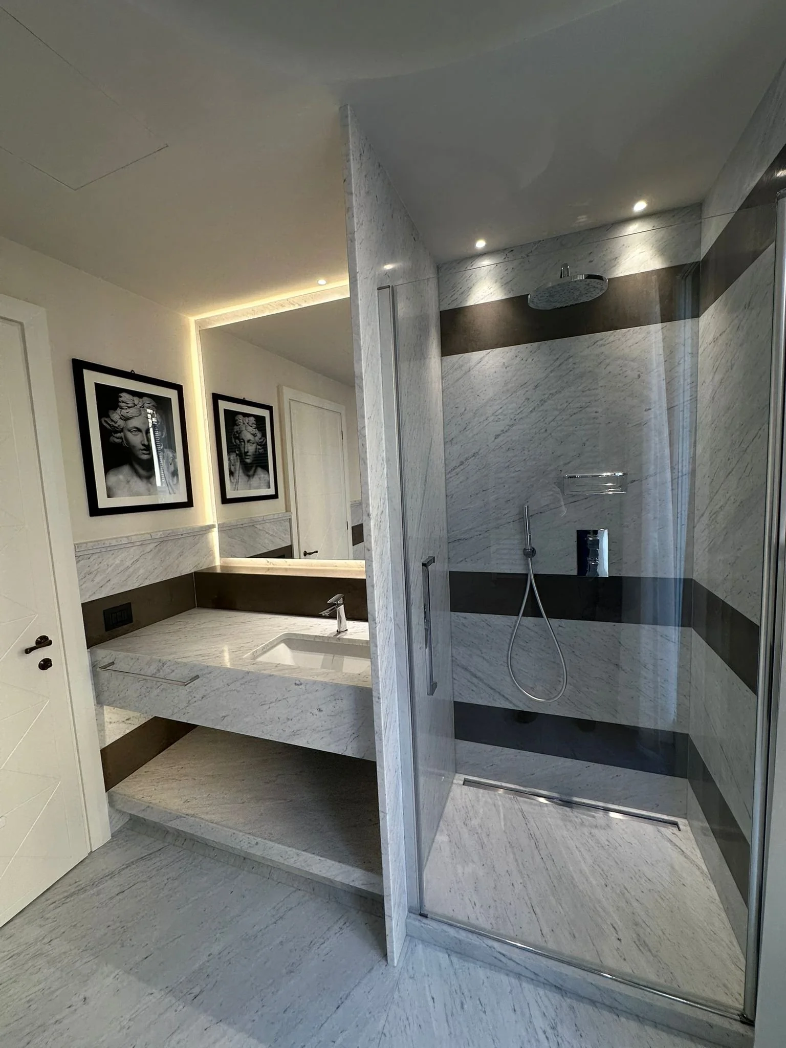 Modern bathroom with marble walls and floors, featuring a walk-in shower with a rain shower head and a handheld shower. The bathroom has artwork of classical sculptures on the wall and minimalist lighting.