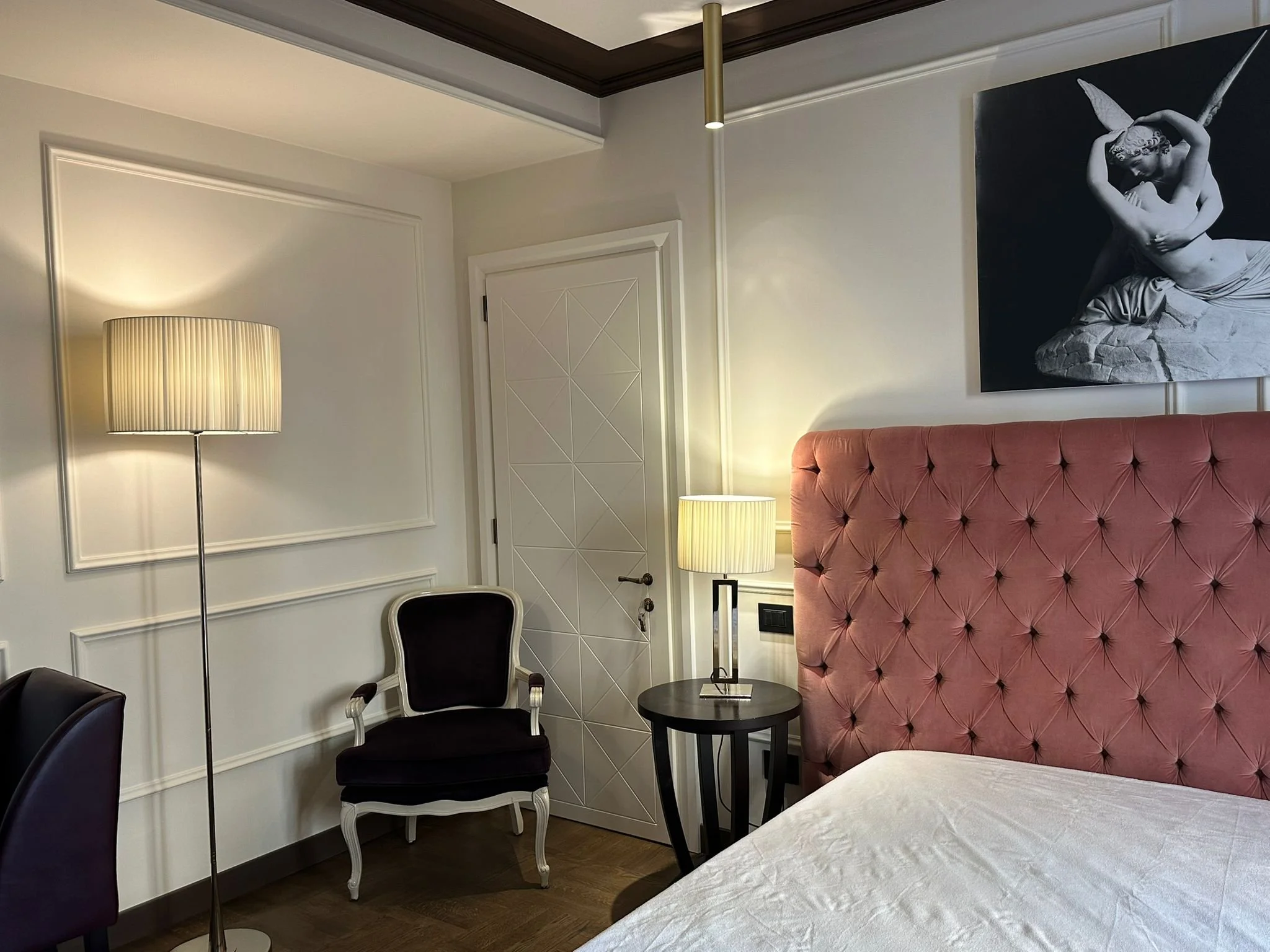 A corner of a bedroom with a pink tufted headboard, a black side table with a lamp, a black and white classical sculpture painting on the wall, a standing lamp, a black chair, and a door with geometric panel design.