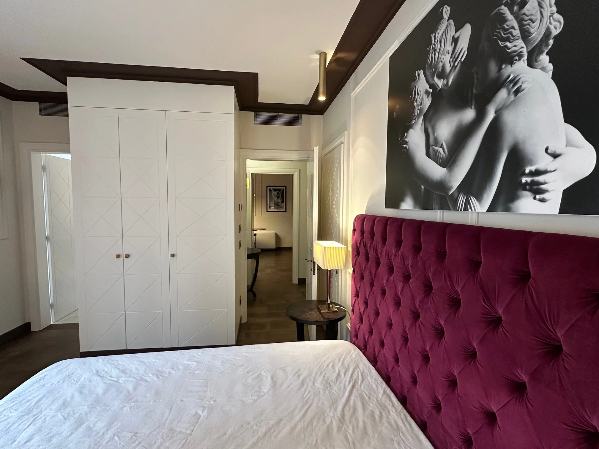 A hotel room with a large black-and-white mural of a classical sculpture of two lovers embracing on the wall behind a plush, deep red velvet headboard. There is a nightstand with a lamp, a small round table, and part of a white bed with a grey blanke