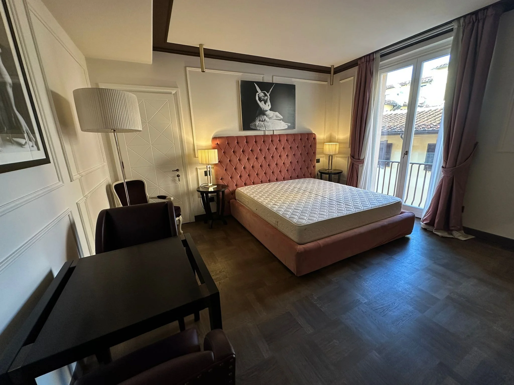 A bedroom with a pink upholstered headboard, nightstands with lamps, a wall art of a stylized woman, large window with curtains, and a black chair.