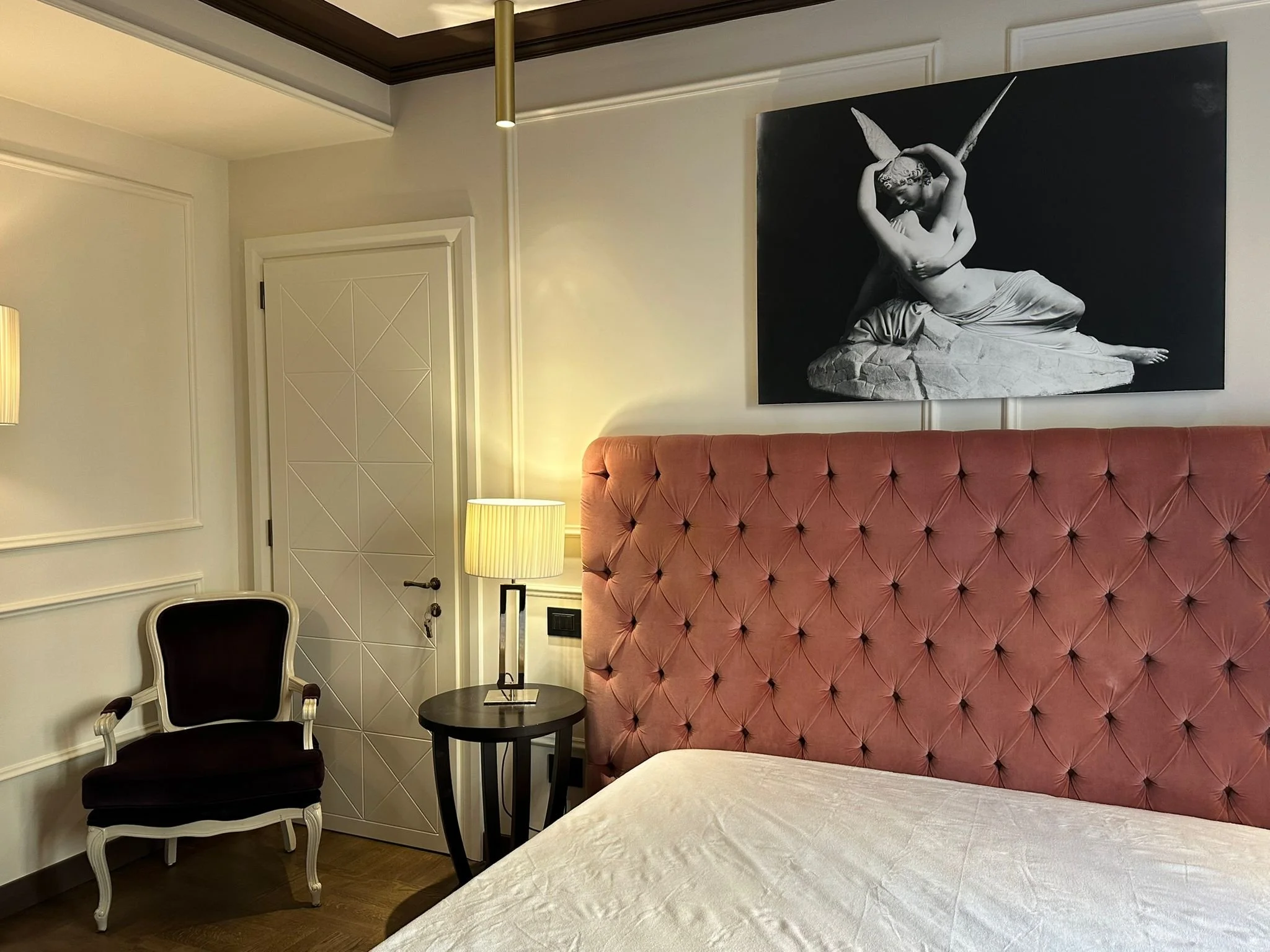 A hotel room corner with a black and white painting of a classical statue, a pink tufted headboard, a small black side table with a lamp, a black and white chair, and a cream-colored bedspread.