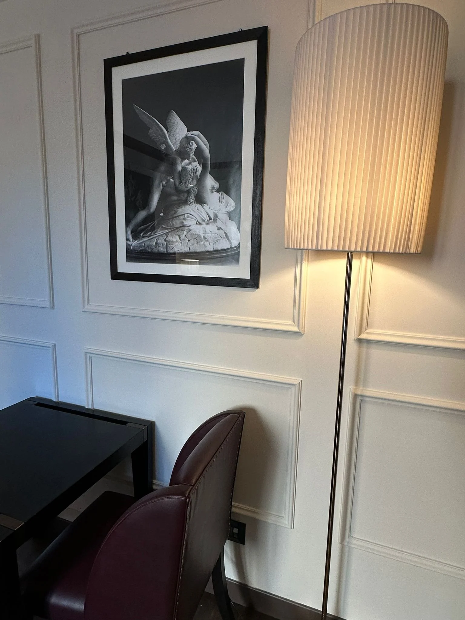 Interior of a room with a black table, maroon chairs, a framed monochrome art print of a mythological figure with butterfly wings, a tall floor lamp with a large shade, white paneled walls.