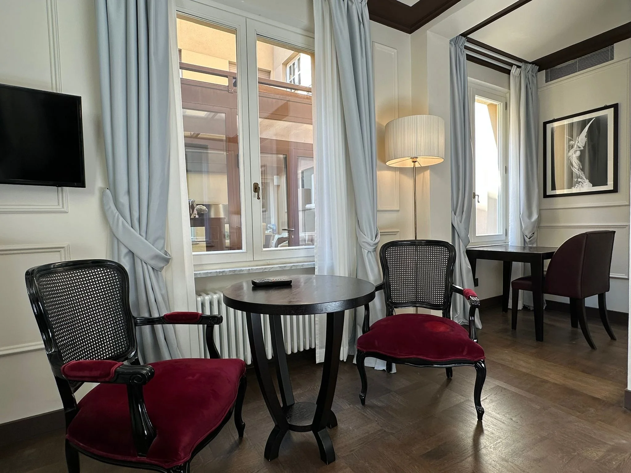 A room with large windows with white curtains, a table with chairs with red velvet cushions, a picture on the wall, a floor lamp, and a television mounted on the wall.