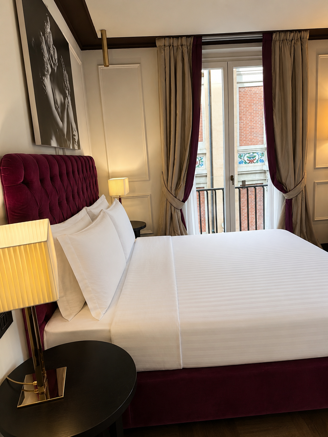 A hotel room with a large bed with white bedding and a deep red tufted headboard. Two bedside tables with yellow lamps flank the bed. There is a large window with beige and deep red curtains, with a view of a brick building outside.