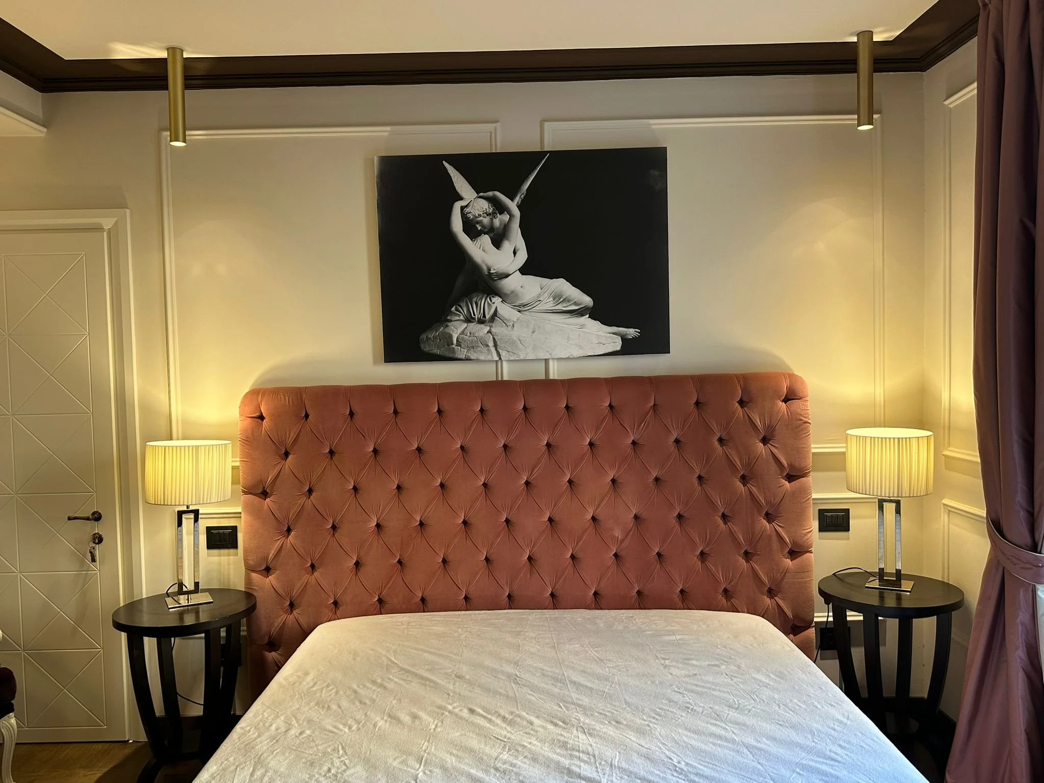 A hotel bedroom with a pink tufted headboard, two side tables with yellow lamps, a white bedspread, and a black and white artwork of a woman with butterfly wings on the wall above the bed.