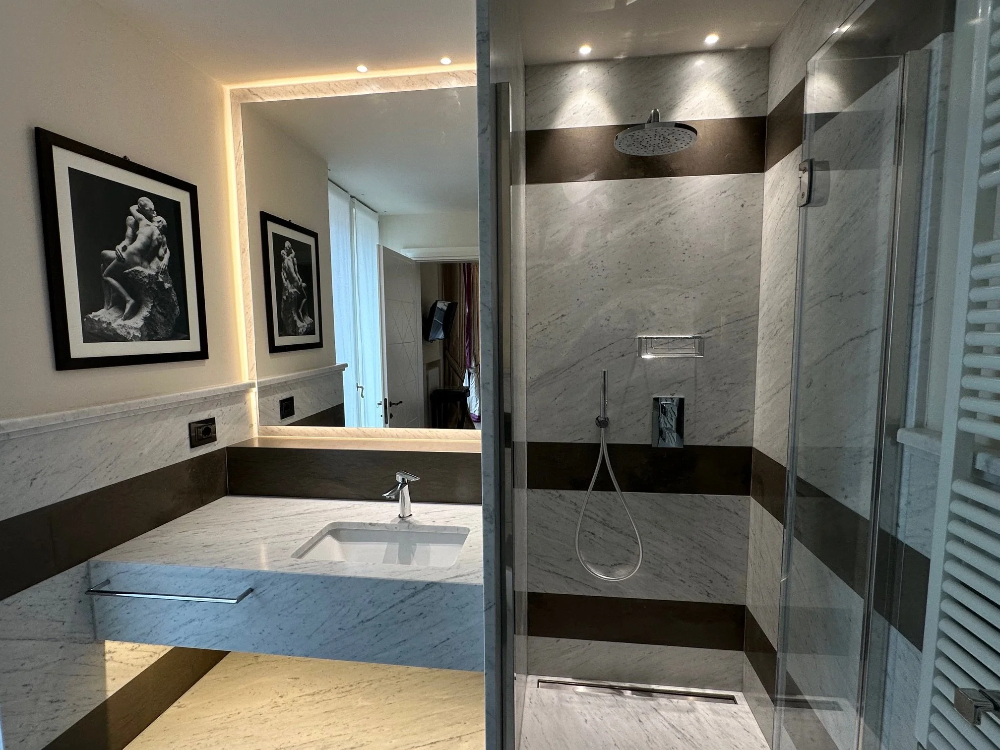 A modern bathroom with a marble shower and a large mirror with built-in lighting. Two framed black-and-white photographs hang on the wall near the mirror.