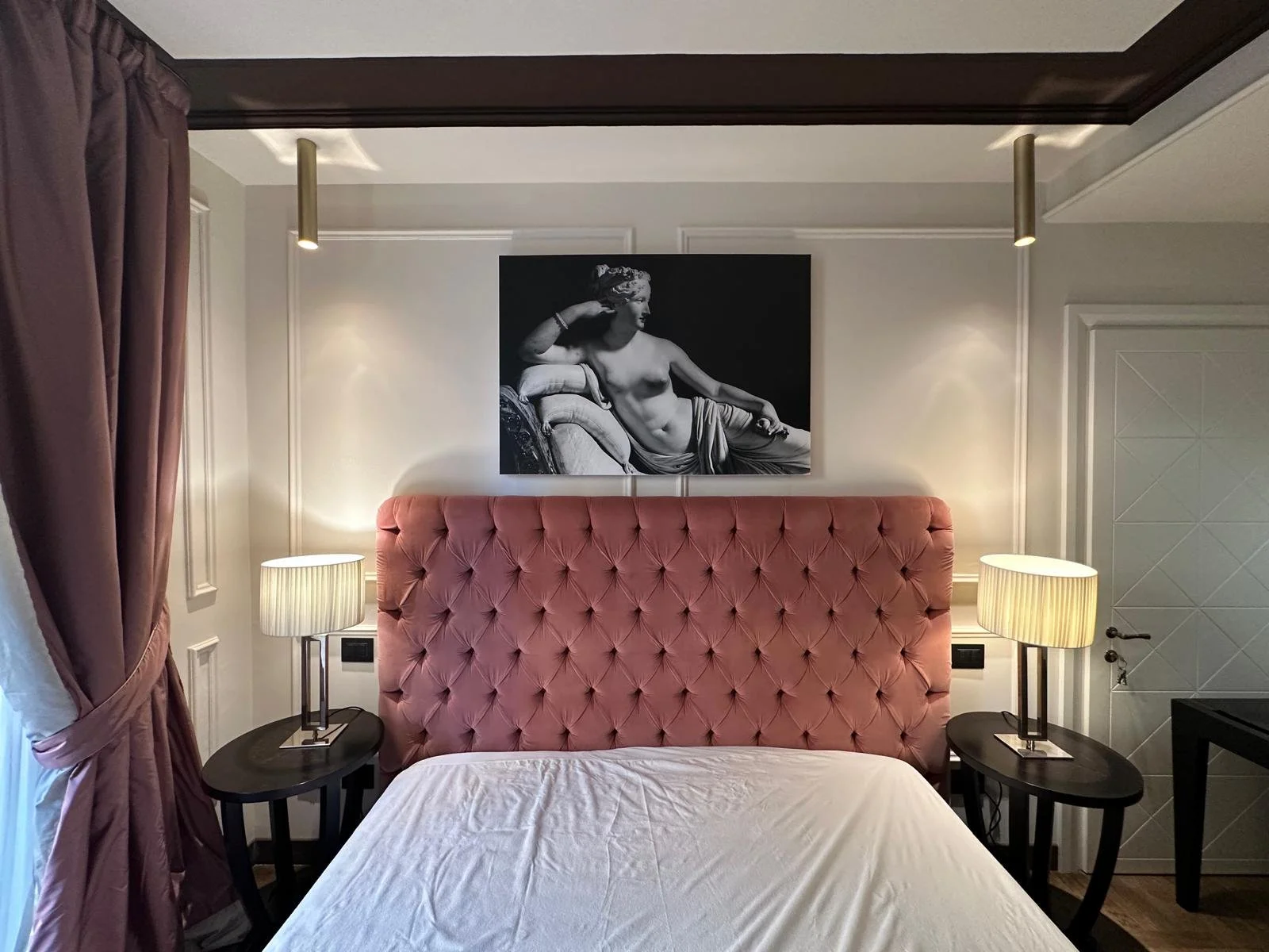 A bedroom with a pink tufted headboard, two black bedside tables with lamps, and a black-and-white art piece of a reclining woman hung above the headboard. There are dark purple curtains on the left side and a white bedspread in the foreground.