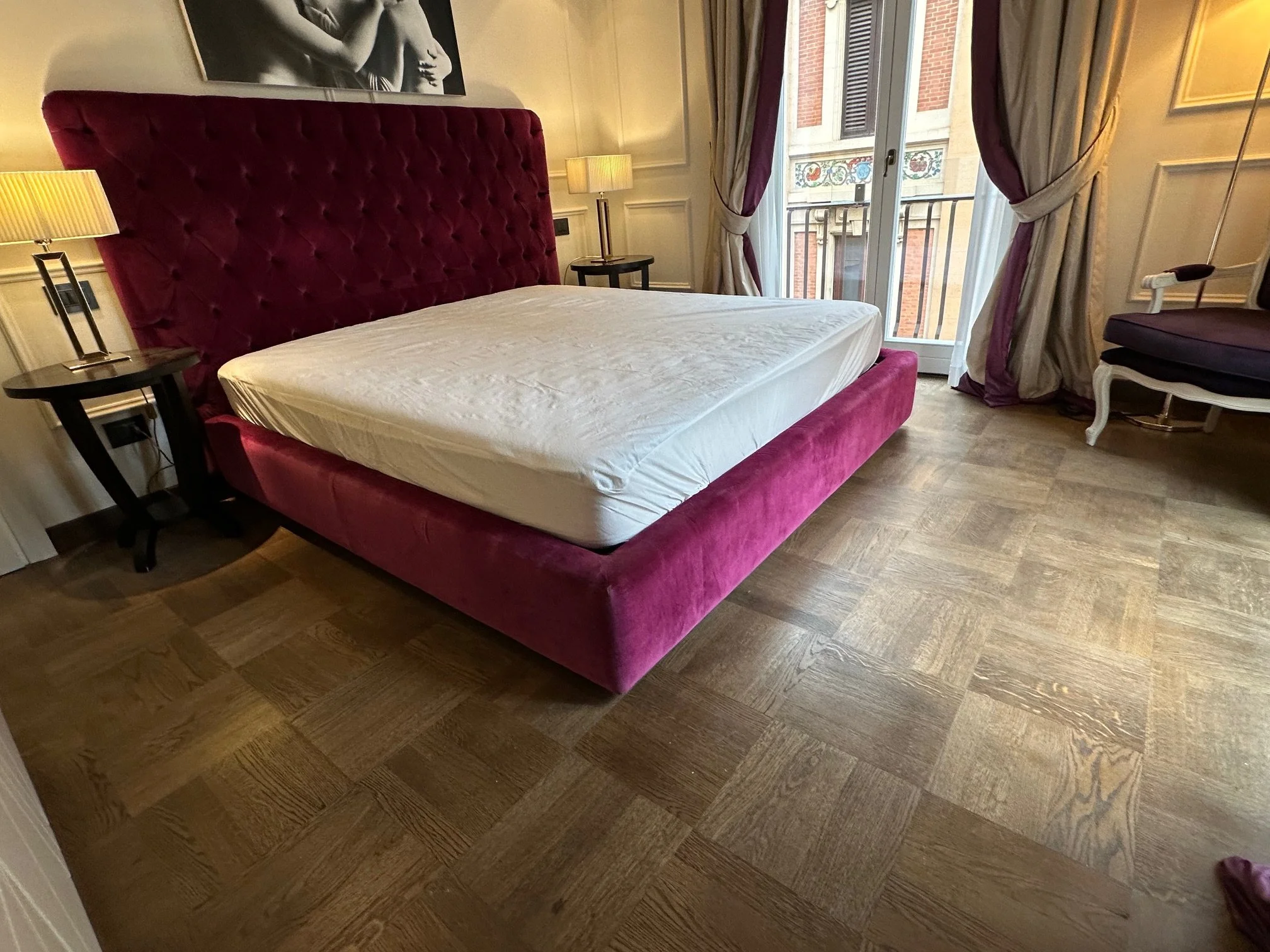 A bedroom with a large pink velvet tufted bed frame and a white mattress. Two nightstands with lamps and a black and white artwork on the wall. Large window with purple and cream curtains, and a view of a building with decorative trim.