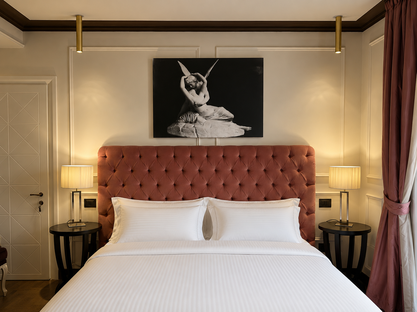 A hotel bedroom with a bed that has a pink, tufted headboard, white bedding, and two white pillows. There are two bedside tables with lamps on each side. Above the bed, a black and white portrait of a woman with wings is mounted on the wall. The room