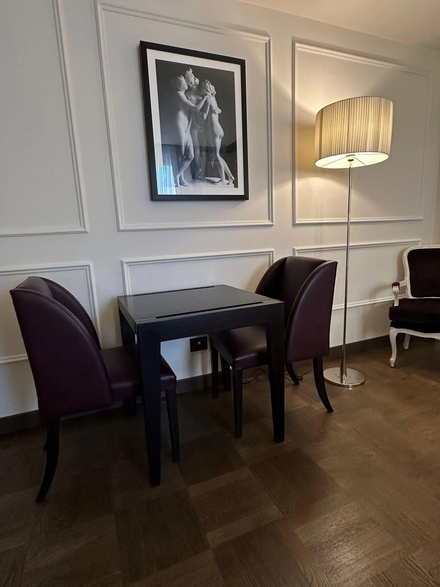 A corner of a room with a black table, four dark red chairs, a standing lamp with a beige, ribbed shade on a metal pole, and a framed black-and-white picture of two human statues on the wall.