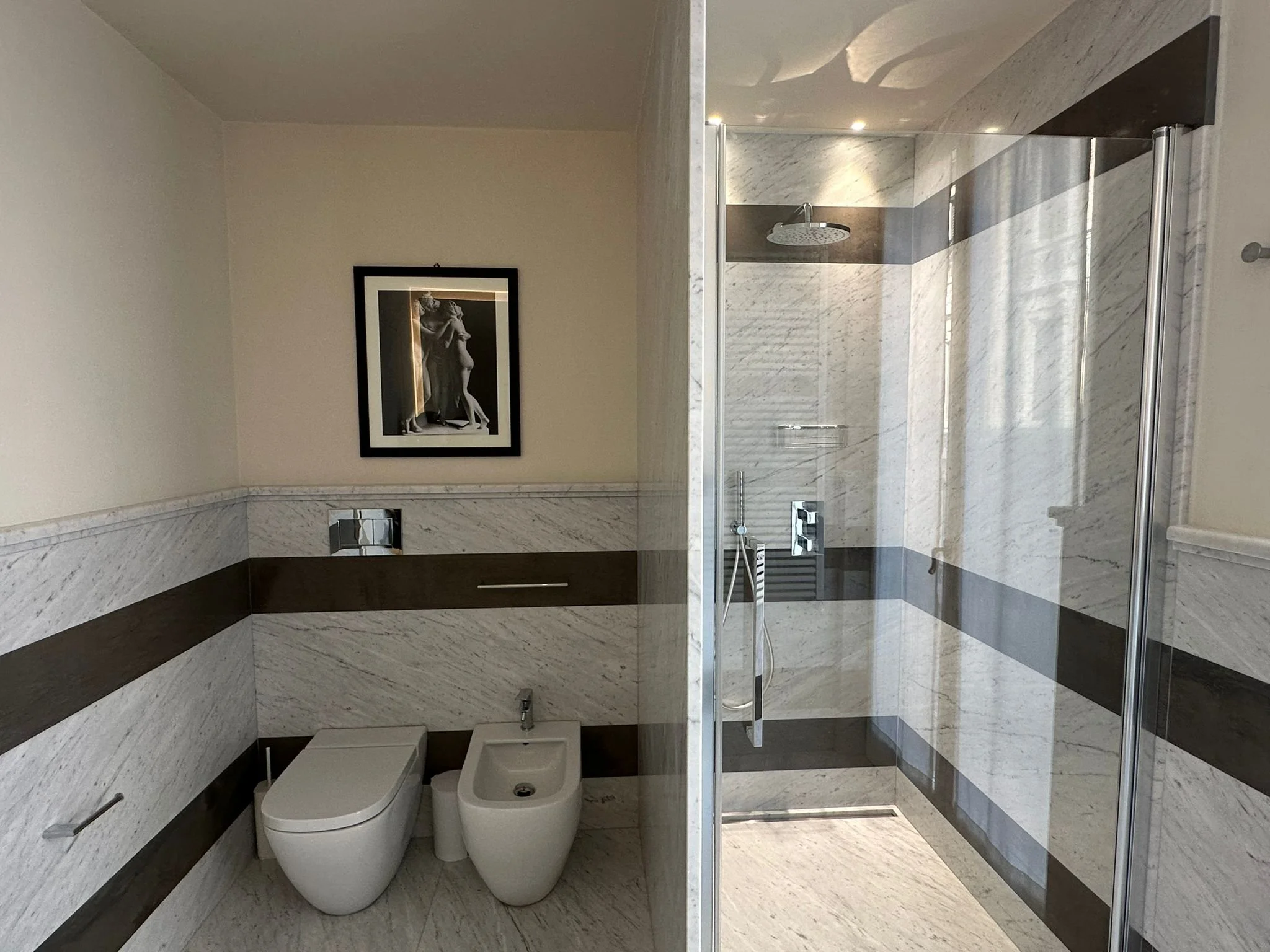 A modern bathroom with marble tiles, a framed black and white photo of a sculpture on the wall, a toilet, a bidet, and a glass-enclosed shower with a rainfall showerhead.
