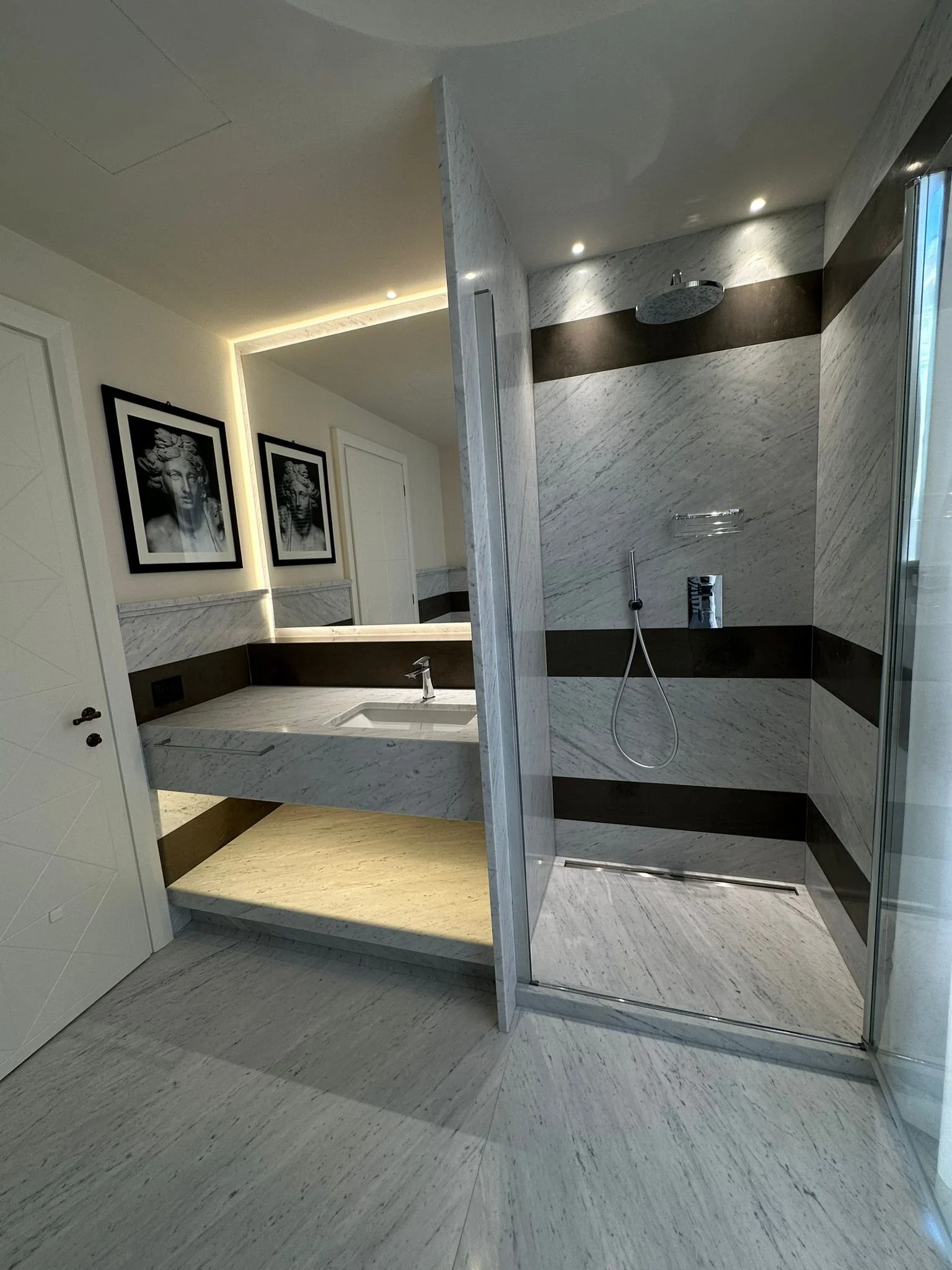 Modern bathroom with marble walls and floors, including a walk-in shower with a rain showerhead, a vanity with a sink, and framed black-and-white artwork on the wall.