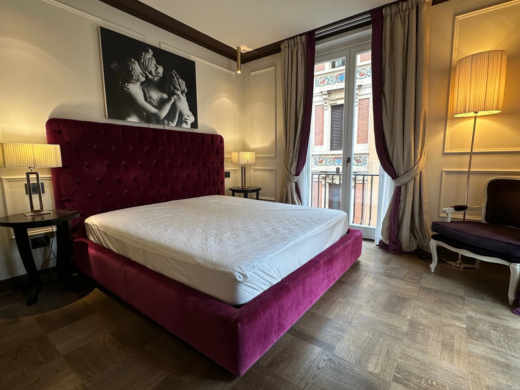 A hotel bedroom with a bed that has a red velvet headboard, two bedside tables with lamps, a large black and white art piece of cherubs on the wall, a window with ornate curtains, and a standing floor lamp.
