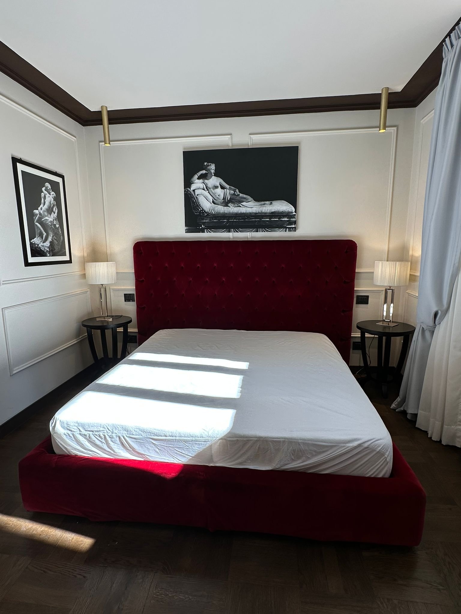 A bedroom with a bed that has a red velvet headboard, a white mattress, and two nightstands with lamps. Two black-and-white framed photographs of classical sculptures hang on the walls, and there are gold-colored ceiling lights.