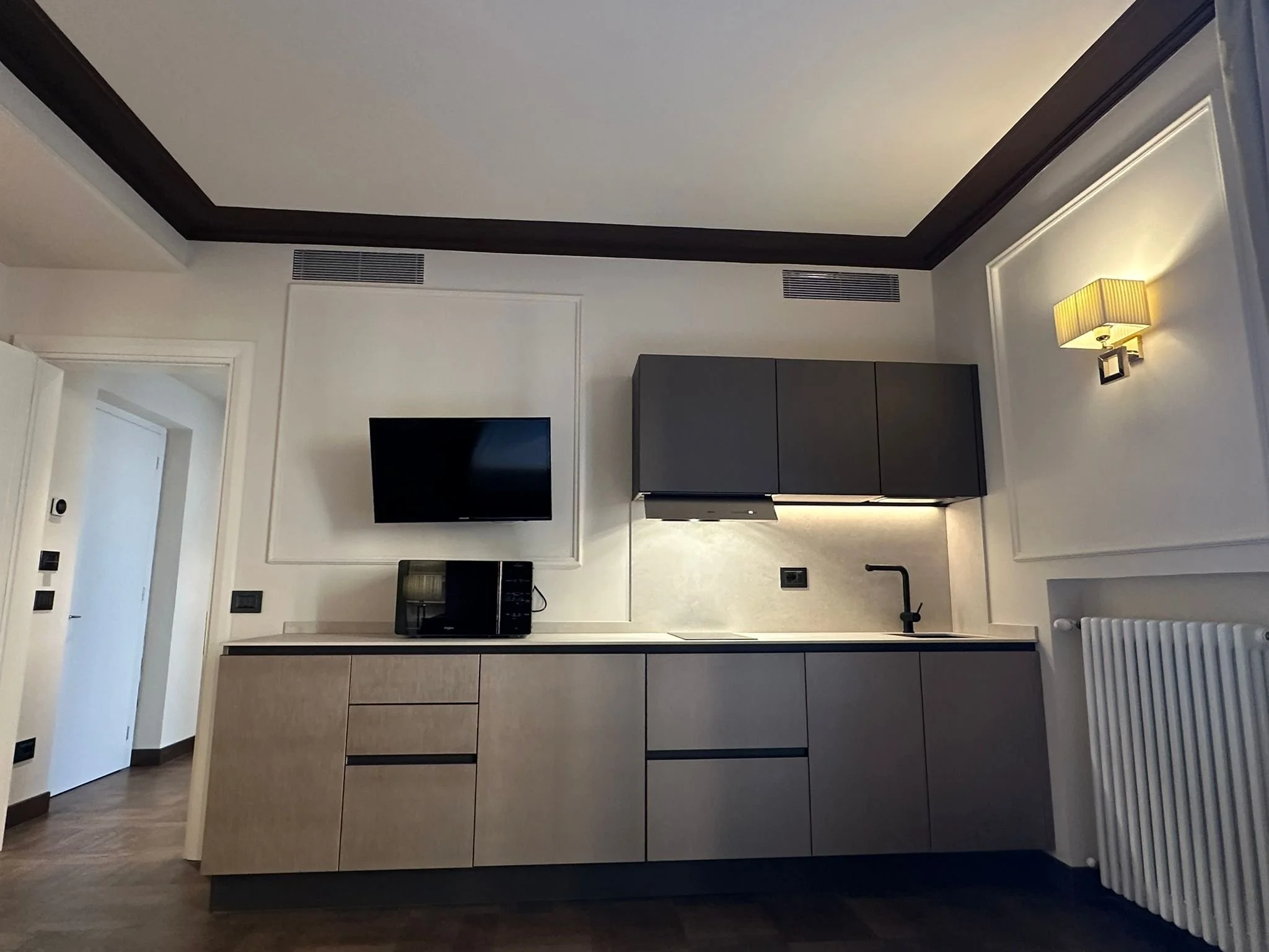 Interior of a modern living room with a flat-screen TV mounted on white wall, dark and light wood cabinets, a wall-mounted lamp, and a radiator.