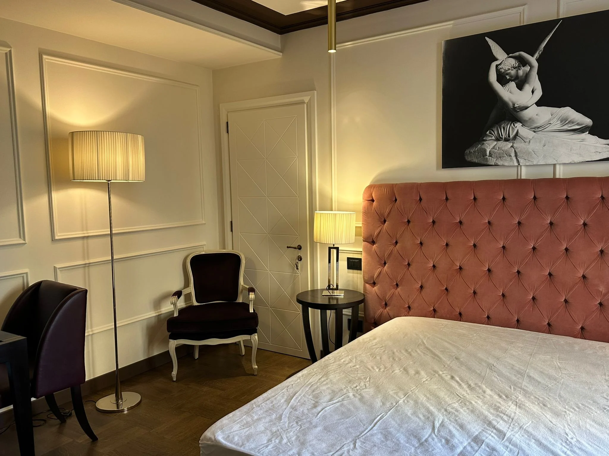 A bedroom corner with part of a bed, a pink tufted headboard, two black chairs, a small round black table, a floor lamp, a door, and a black and white painting of a woman with wings.