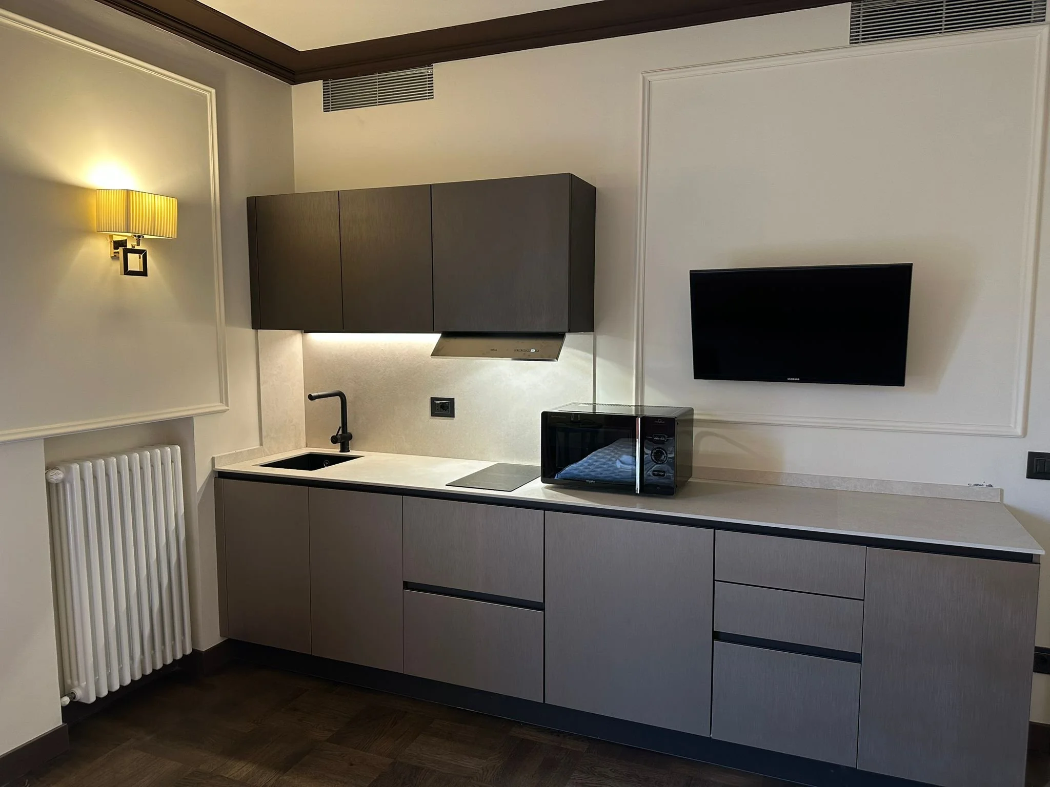 A modern kitchen with gray cabinets, white countertop, a black sink, and a stove. A yellow striped wall lamp is mounted on the wall, and a black flat-screen TV is on the right. The room has cream walls with decorative molding and dark wood flooring.