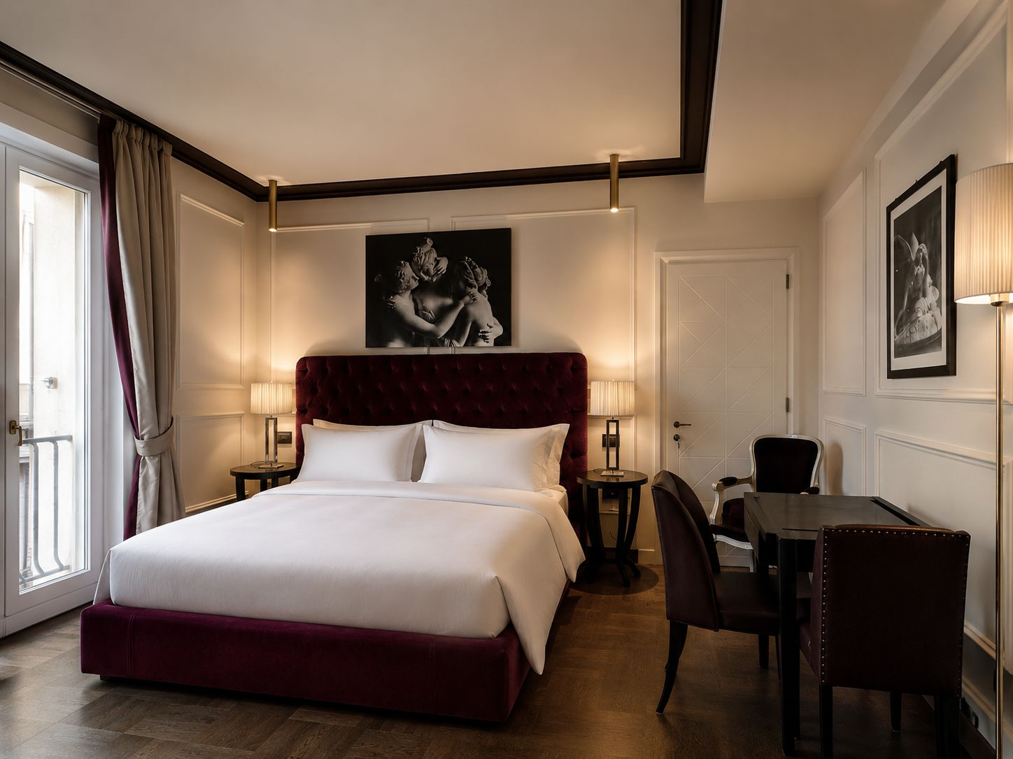 A luxury hotel room with a large bed featuring a red tufted headboard, side tables with lamps, dark wood flooring, artwork above the bed, a small table with chairs, and a balcony door with curtains.