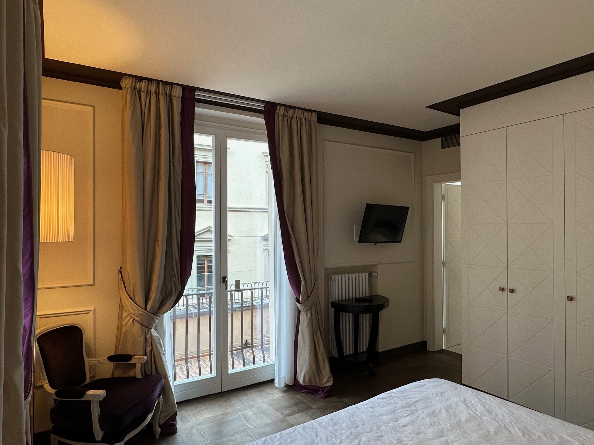A hotel room with a window, curtains, a chair, a wall-mounted TV, and a bed