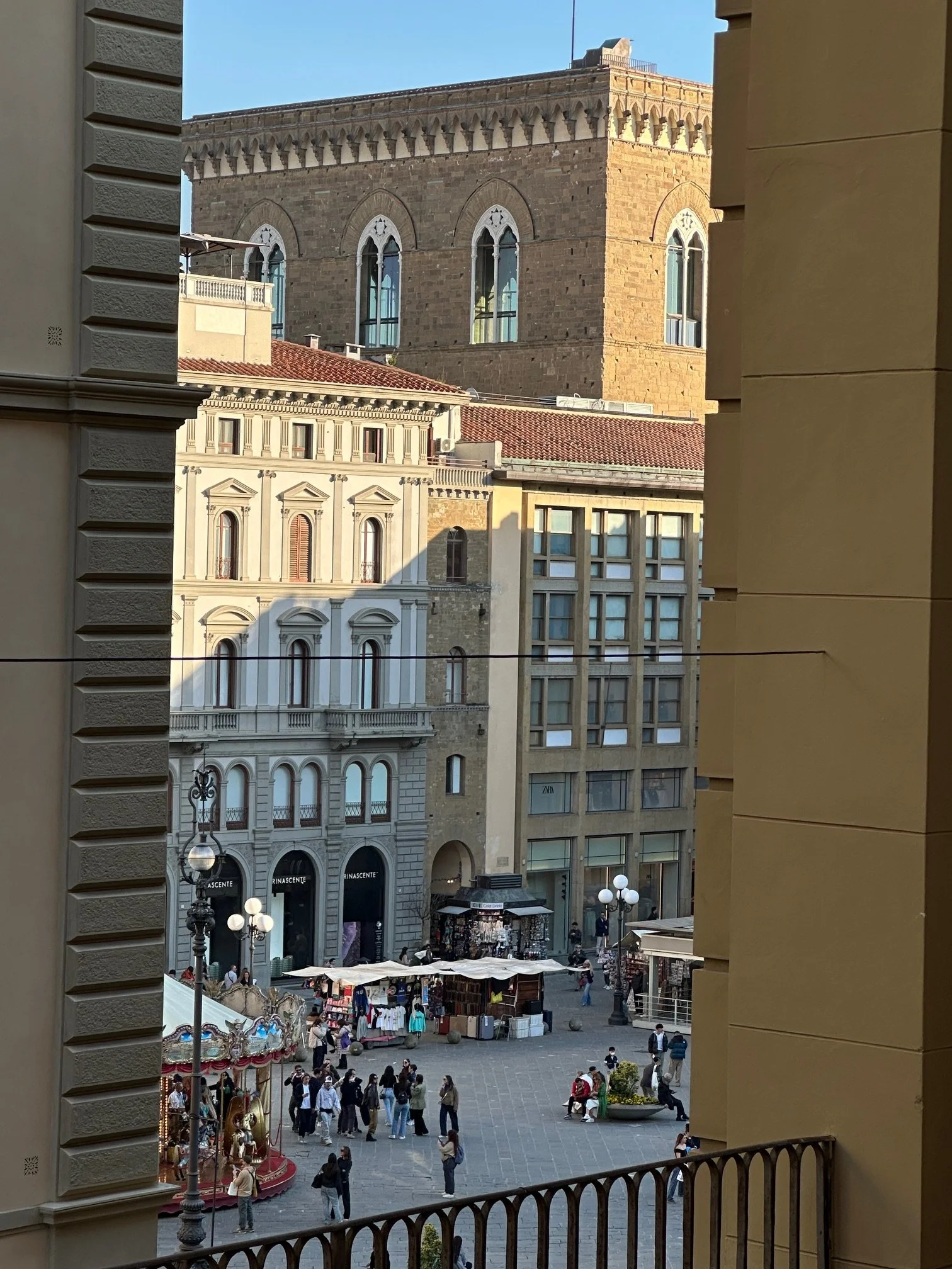 City square viewed from a window with ornate railing, featuring historic and modern buildings, street vendors, and numerous pedestrians.