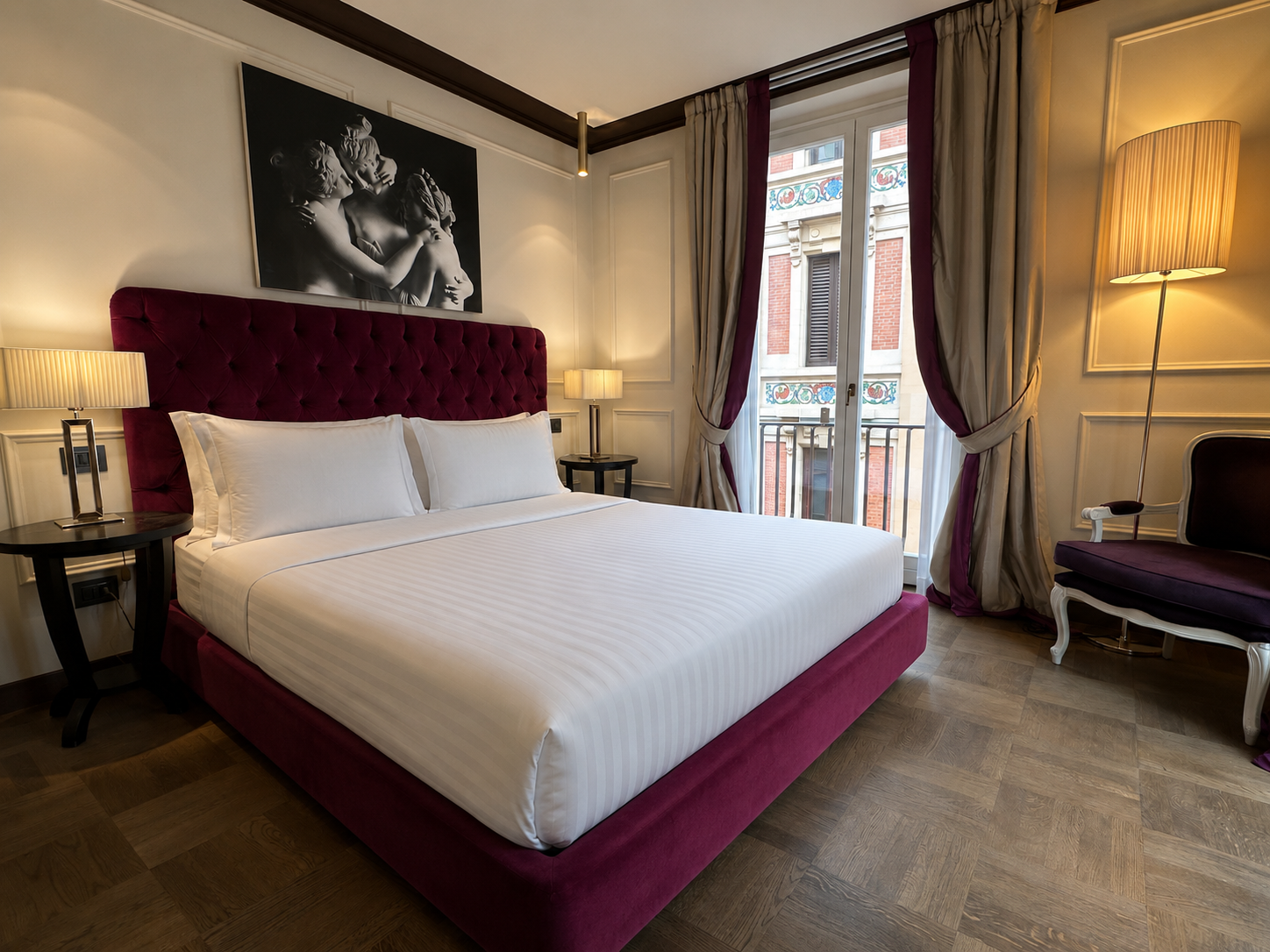 A luxurious hotel room with a large bed featuring a deep red tufted headboard, two nightstands with lamps, a balcony door with curtains, and a black-and-white artwork above the bed. Additional seating includes a vintage-style chair and a tall floor l