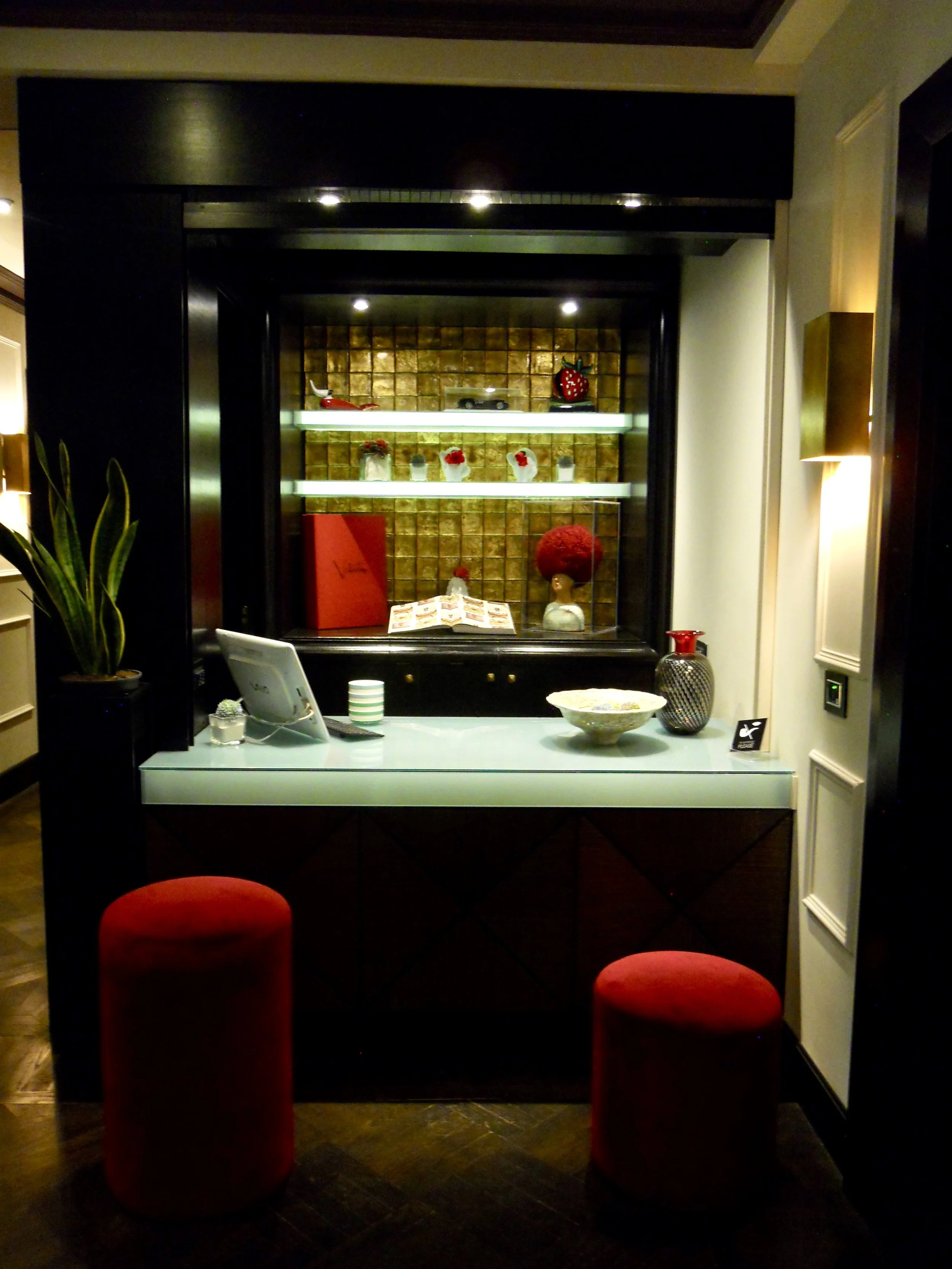 Interior of a modern room with a display cabinet in the background, decorated with a strawberry ornament, several glass jars with flowers or decorations, a large red book, and a small toy car. In the foreground, a glass-top table with a bowl, a strip