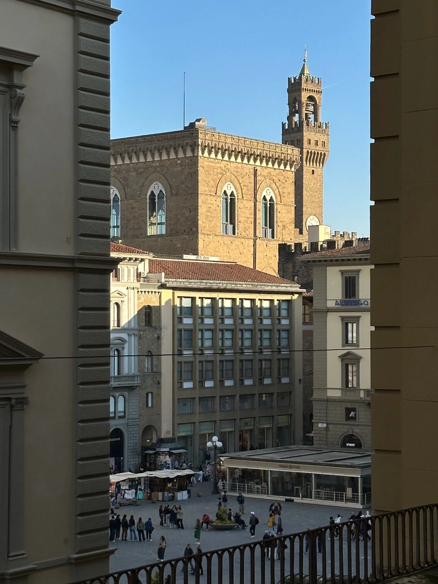 View of a city square with people walking and outdoor market stalls, framed by two buildings with a historic tower in the background under a clear blue sky.