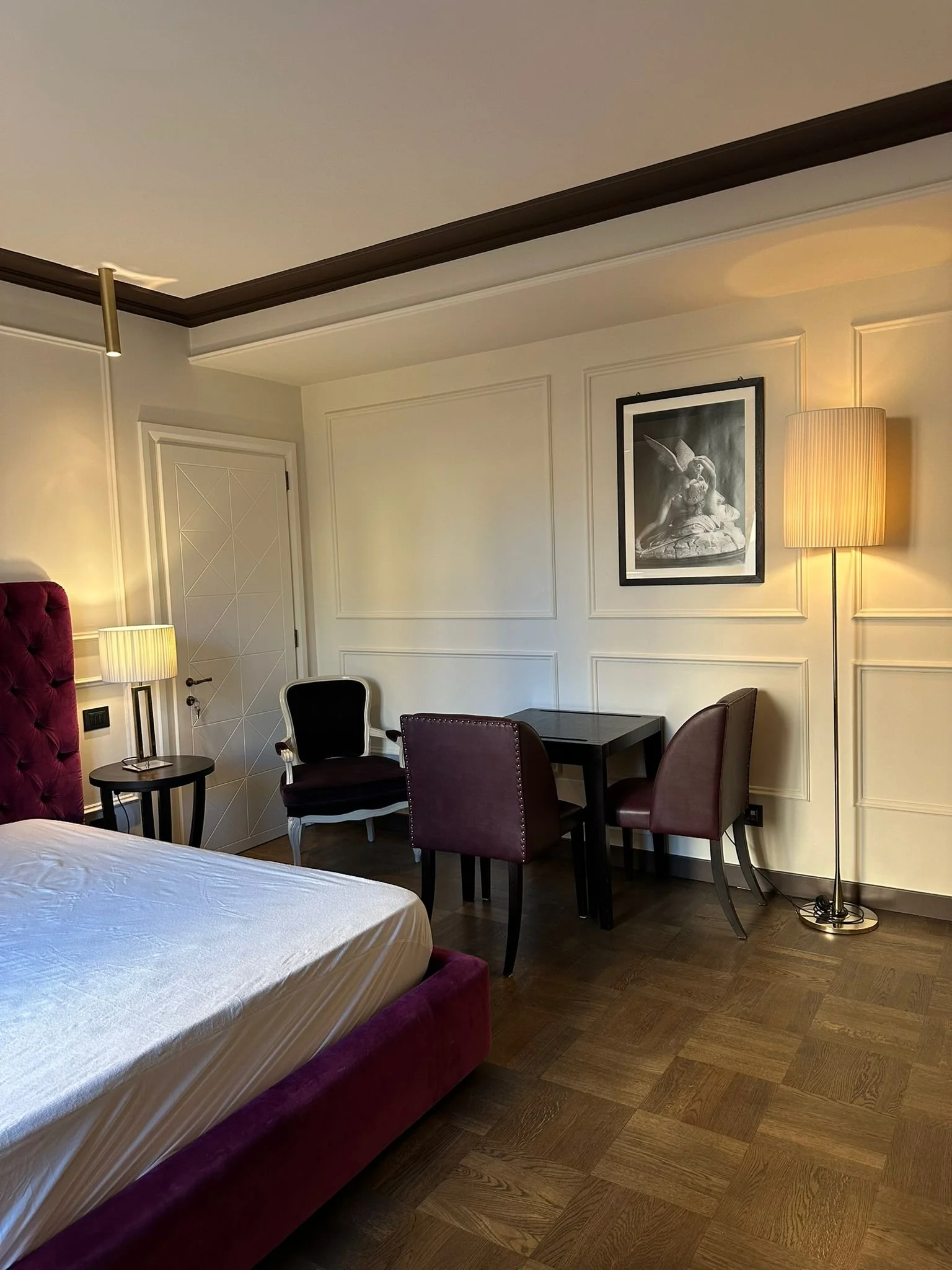 A cozy hotel room corner with a bed, a small round side table, a purple armchair, and a black table with four purple chairs. The wall features decorative molding, a framed black and white art piece of a woman with rabbit ears, a tall lamp with a beig