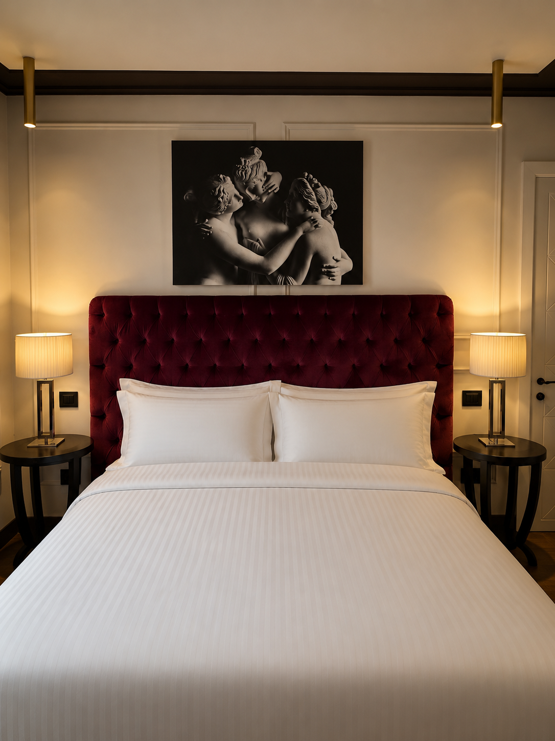 A hotel bedroom with a white bed, large red tufted headboard, two matching nightstands with lamps, and a black and white framed artwork of three classical sculptures on the wall.