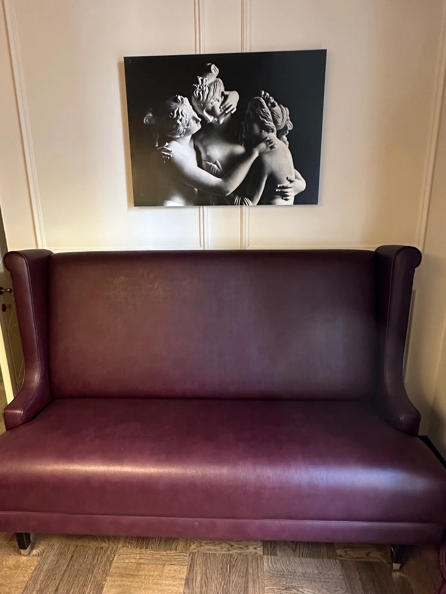 A maroon upholstered bench placed in front of a cream-colored wall with decorative paneling. Above the bench hangs a black-and-white photographic print of three classical-style sculptures depicting embracing figures.