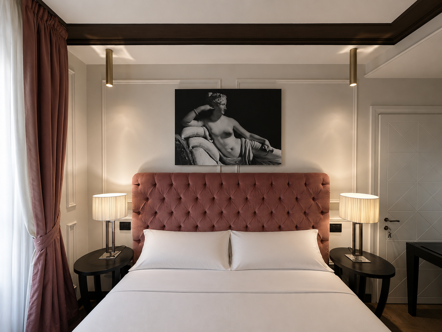 A hotel bedroom with a pink tufted headboard, white bed linens, two bedside lamps, a black and white statue picture above the bed, and pink curtains.