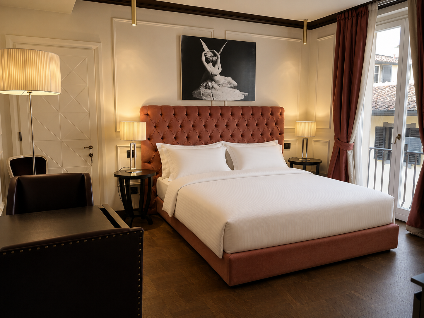 A bedroom with a large bed and a salmon-colored tufted headboard, two bedside lamps, a painting above the bed, a large window with floor-length curtains, a small round table, a chair, and a door.