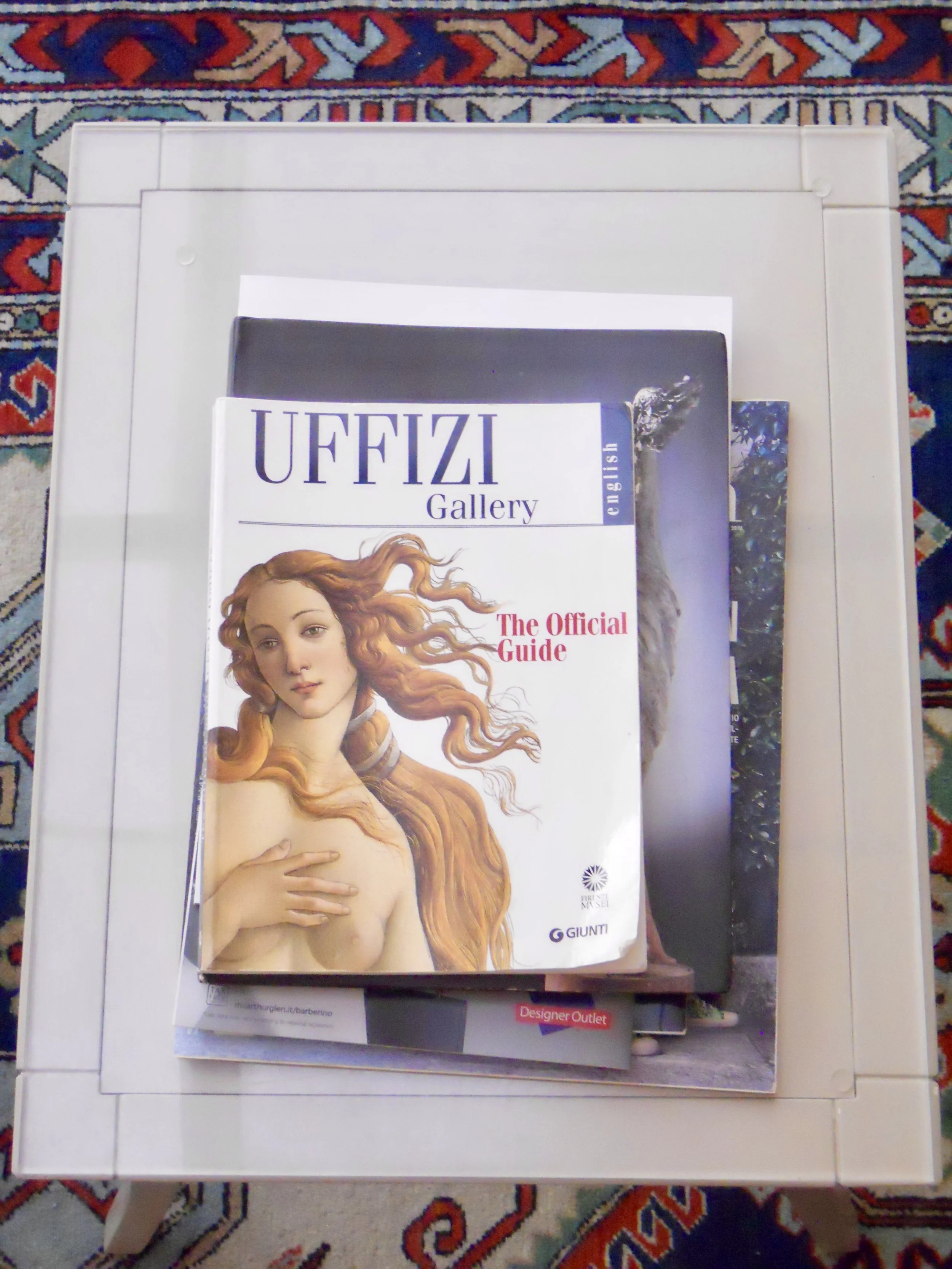 A magazine titled 'UFFIZI Gallery' with a cover image of a woman with long, curly, reddish-blonde hair, looking at the camera with her hand over her chest, laid over a book or brochure on a white table or surface. The background is a patterned orient