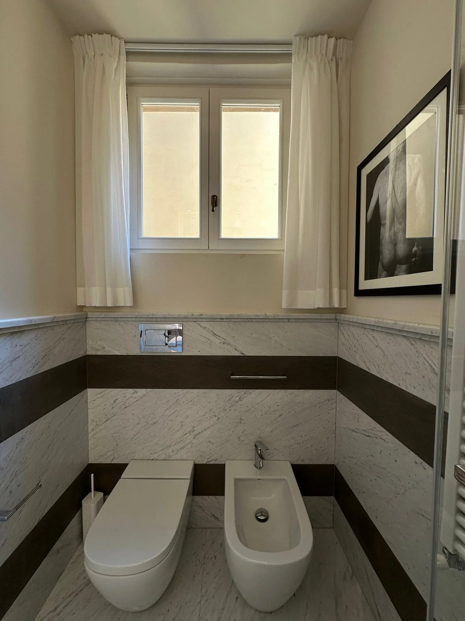 A modern bathroom with a window, white curtains, and marble walls featuring two toilets, a bidet, and a framed black-and-white photograph of a smiling man on the wall.