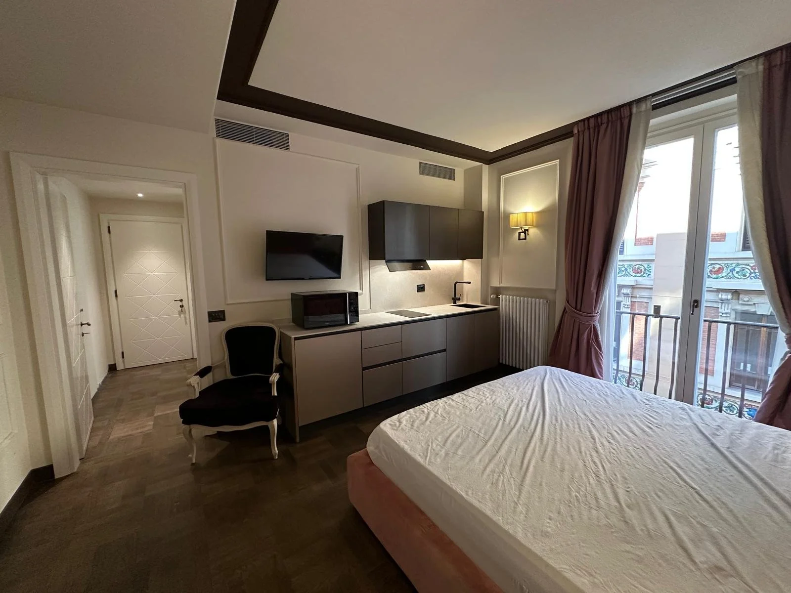 Interior of a modern hotel room with a bed, a kitchenette, a chair, and large windows with pink curtains, overlooking a building with ornate architecture.