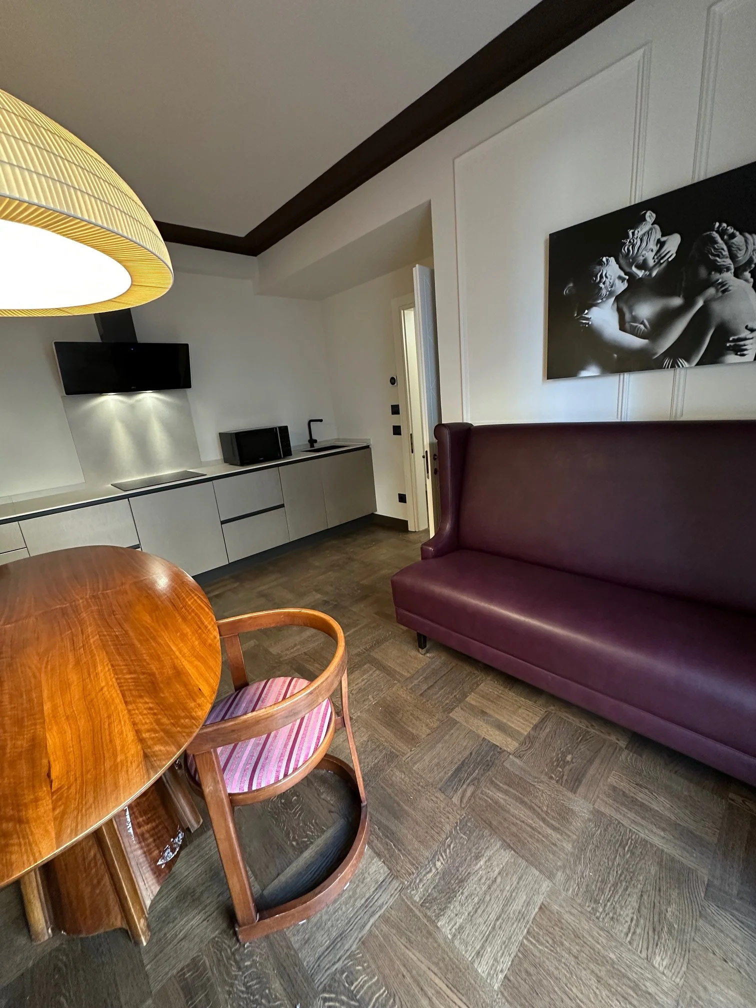 A room with a wooden dining table, pink-striped chairs, a maroon sofa, a wall-mounted TV, a kitchen area with a microwave and sink, a large black and white photograph of classical sculptures, and dark wood ceiling trim.